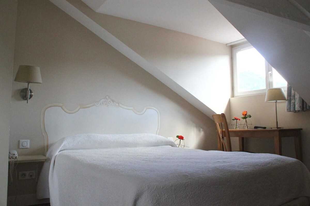 A bedroom with a bed, a nightstand, a desk with a chair, a lamp, and a window.