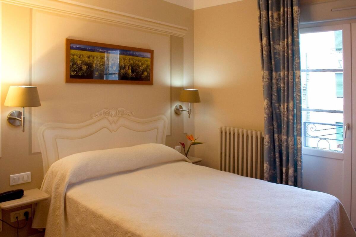 A hotel room with a bed, two bedside lamps, a painting on the wall, a radiator, and a window with curtains.