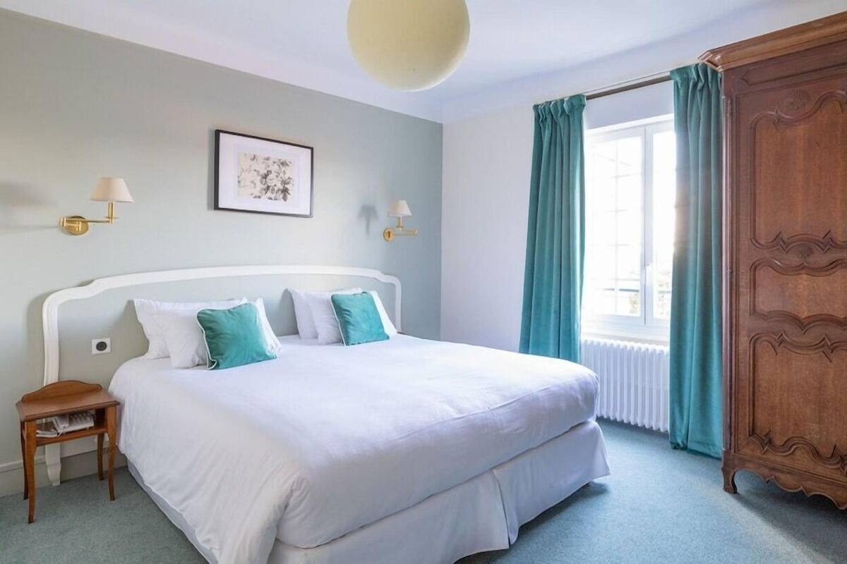 A bedroom with a bed, a nightstand, a wooden wardrobe, a window with curtains, and wall-mounted lamps.
