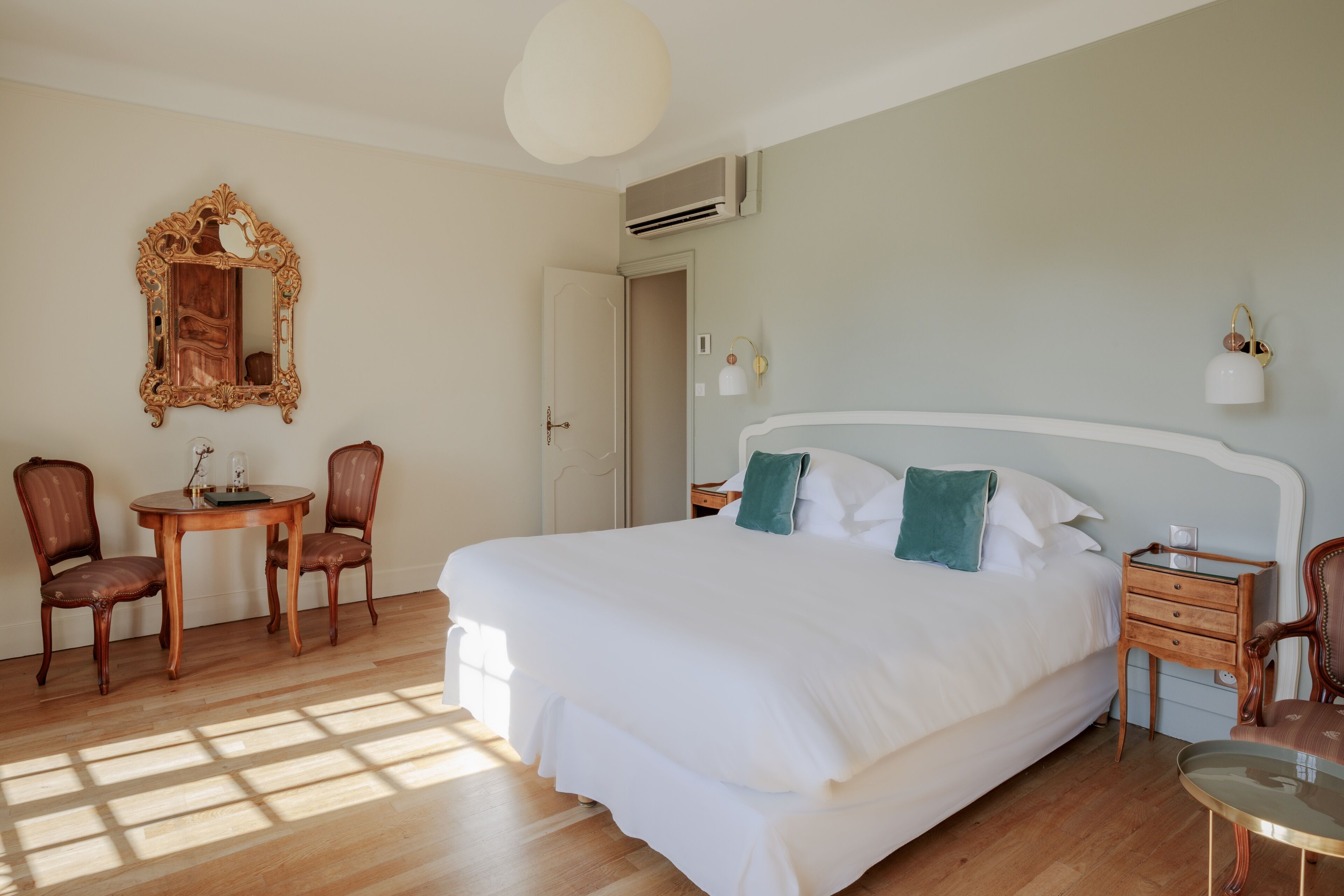 A bedroom with a large bed, a wooden table with chairs, a mirror, and a bedside table with a lamp.