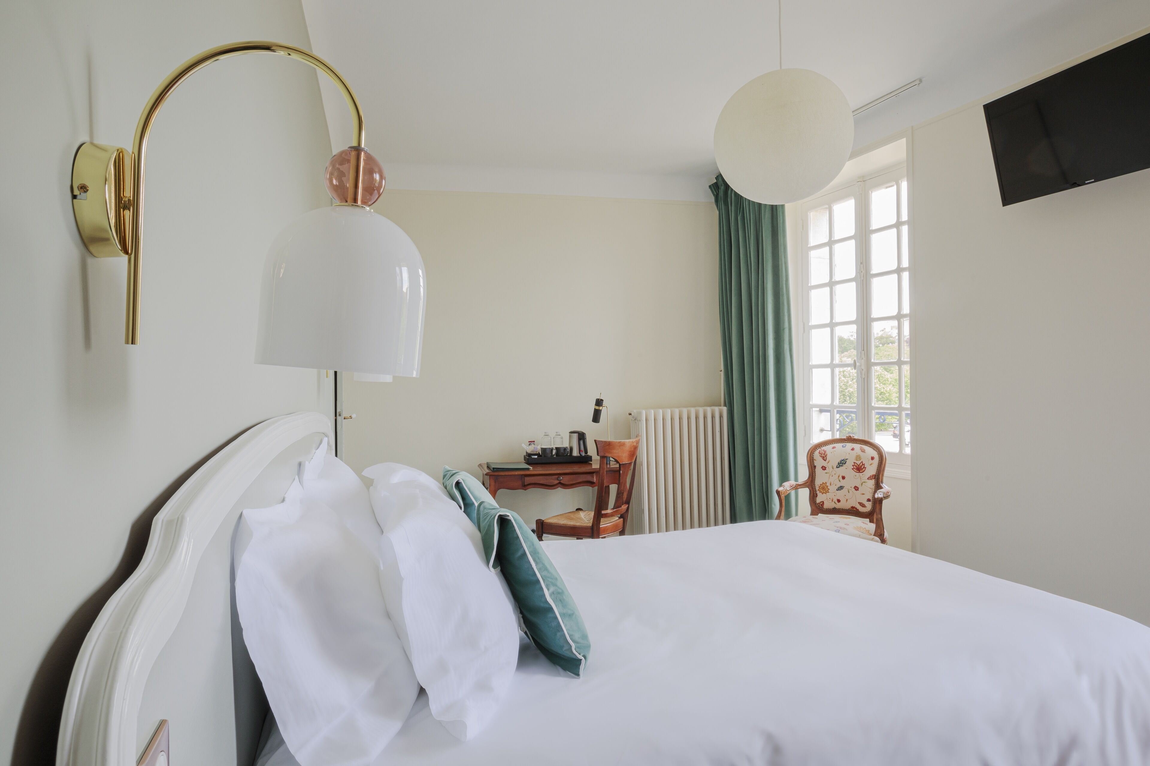 Ein Hotelzimmer mit Bett, Schreibtisch, Stuhl, Lampe und einem fenster mit Vorhängen.