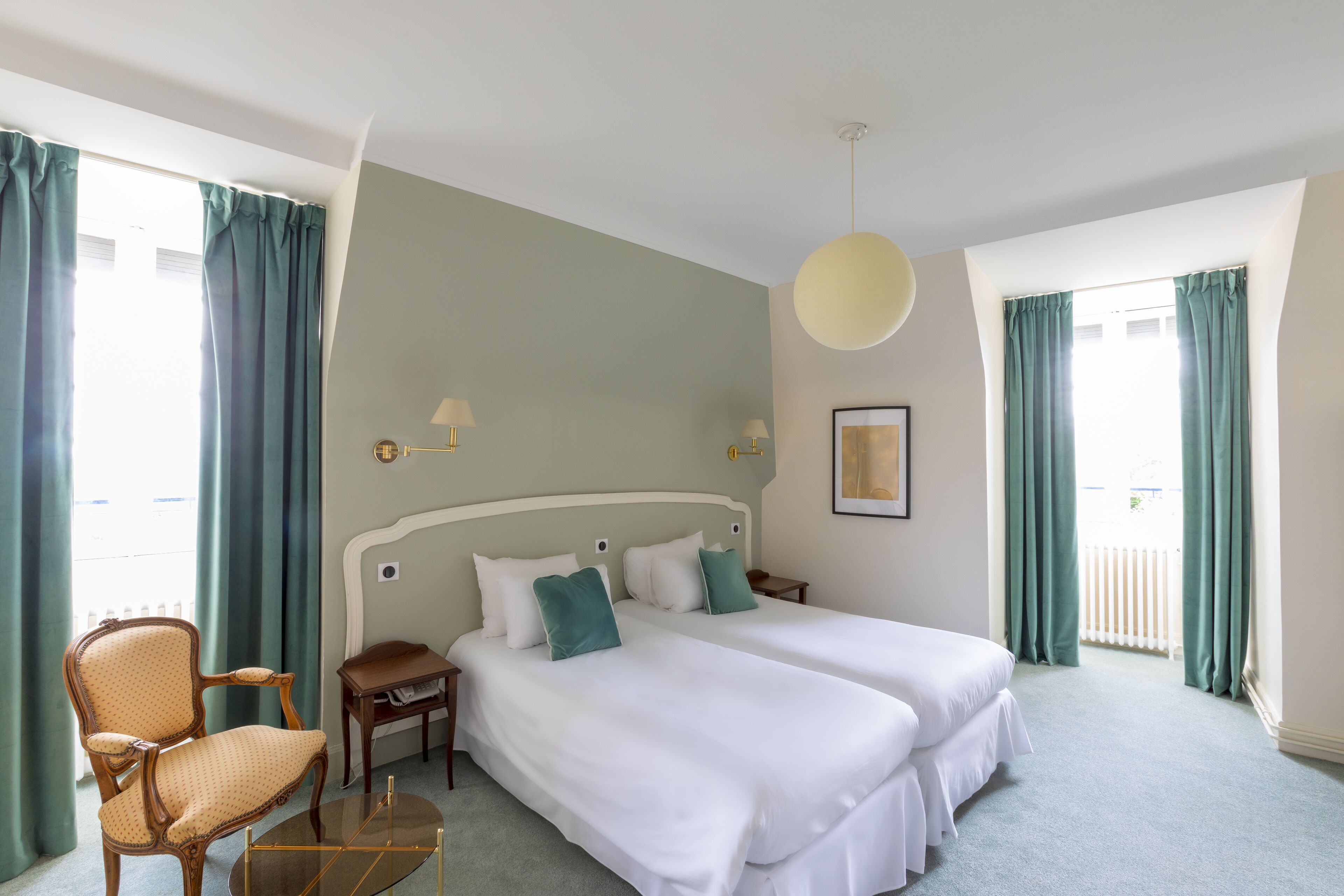 A hotel room with two beds, a chair, a small table, and a hanging light fixture.