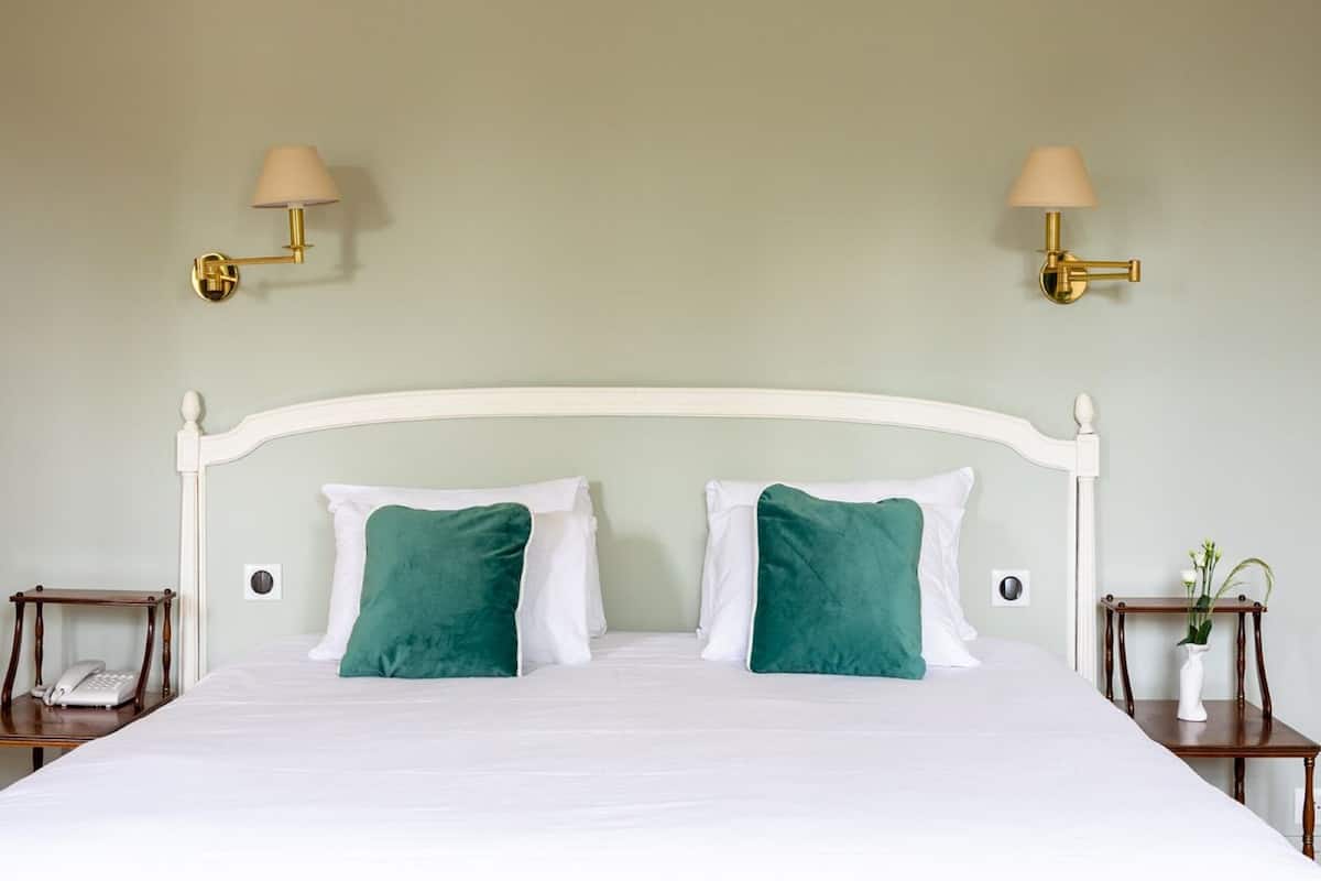A neatly made bed with white linens and two green pillows, flanked by two bedside tables with lamps and a vase of flowers.