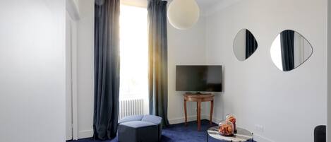 Una sala de estar moderna con alfombra azul, sofá gris, un taburete redondo, una pequeña mesita de noche con un jarrón y un televisor de pantalla plana sobre un mueble de madera.