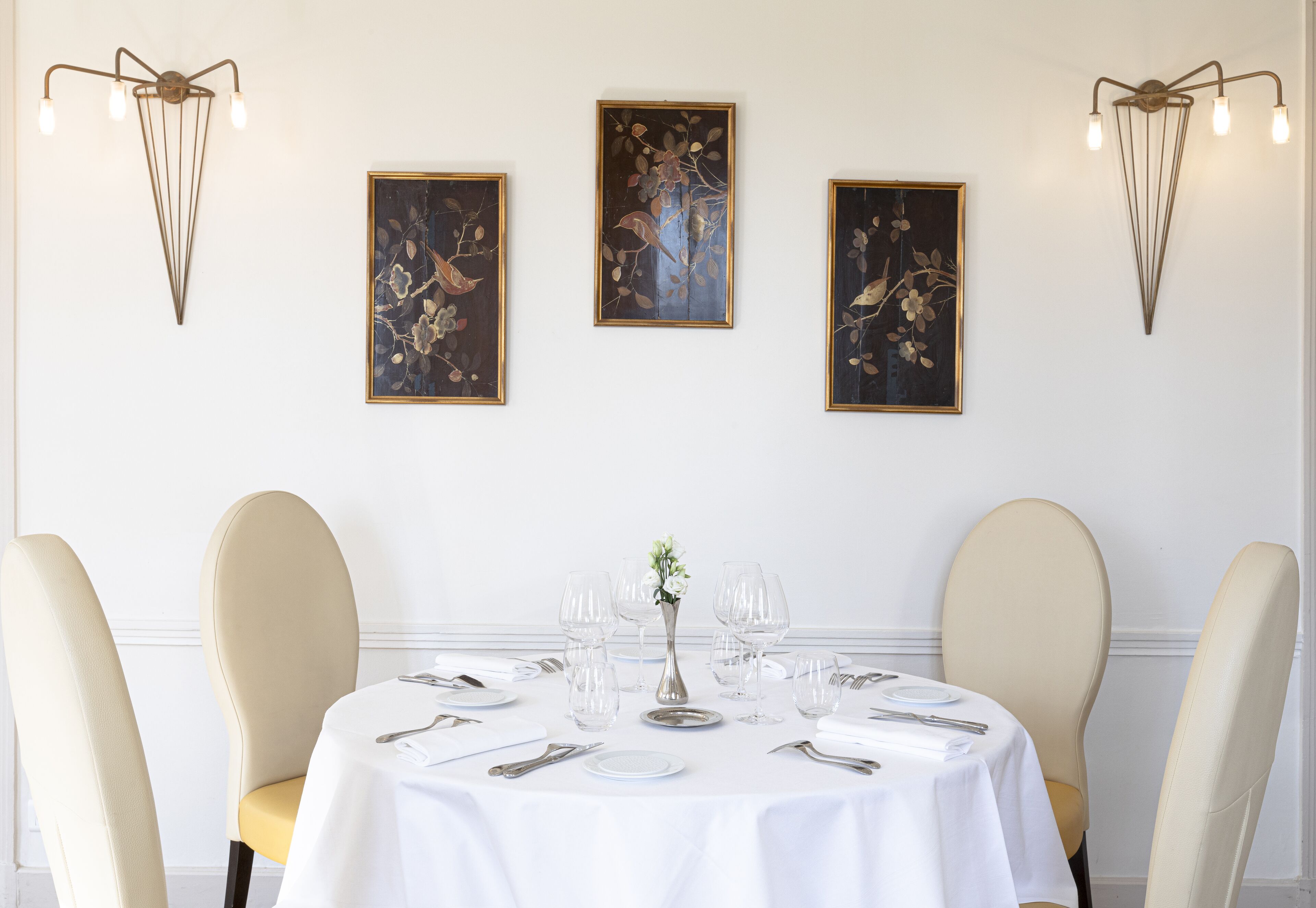 Een eettafel gedekt voor zes personen, met een witte tafelkleed, zes stoelen en een vaas met bloemen. Drie kunstwerken hangen in lijsten aan de muur boven de tafel.