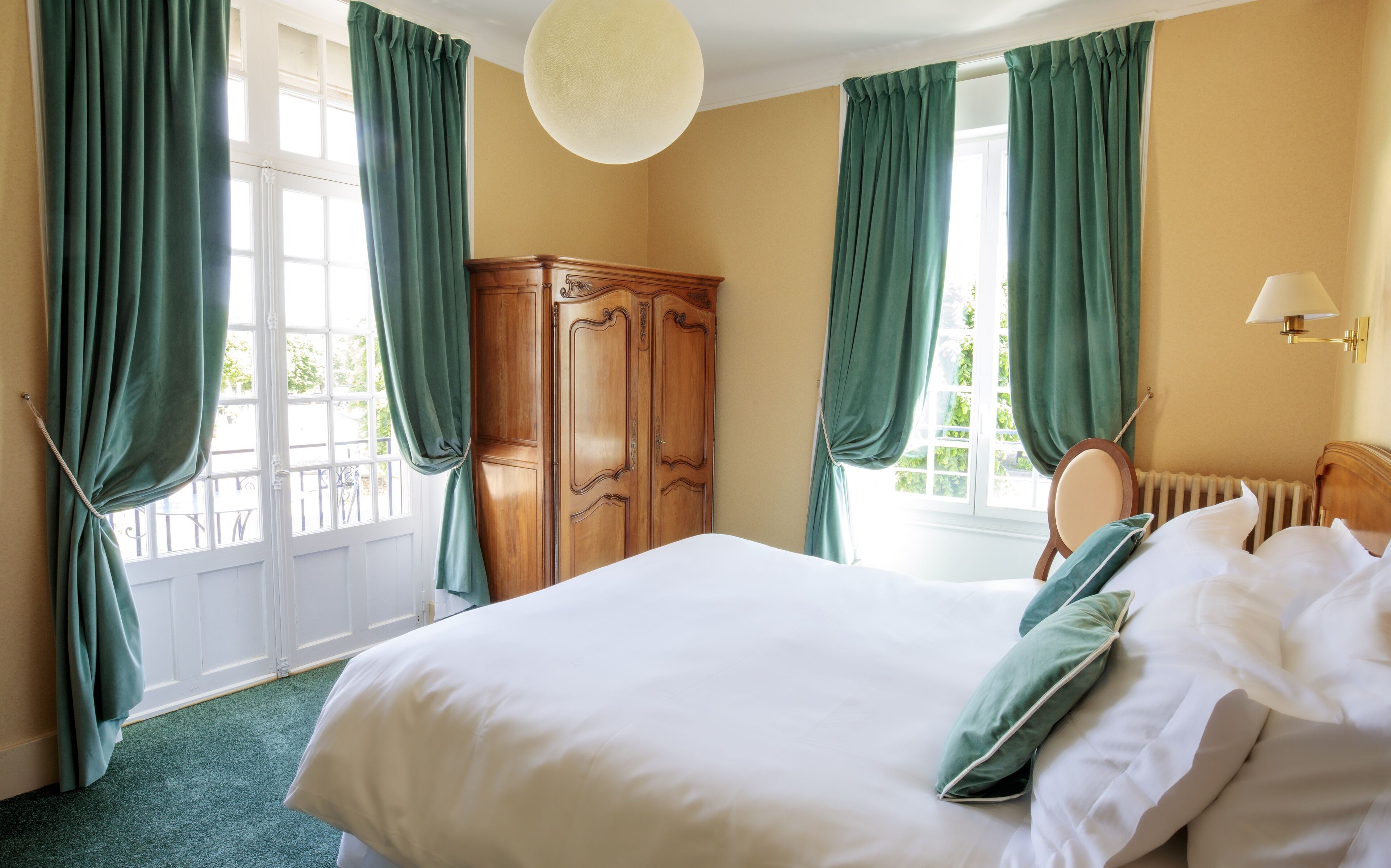 A bedroom with a large bed, green curtains, a wooden wardrobe, and a window with a view.