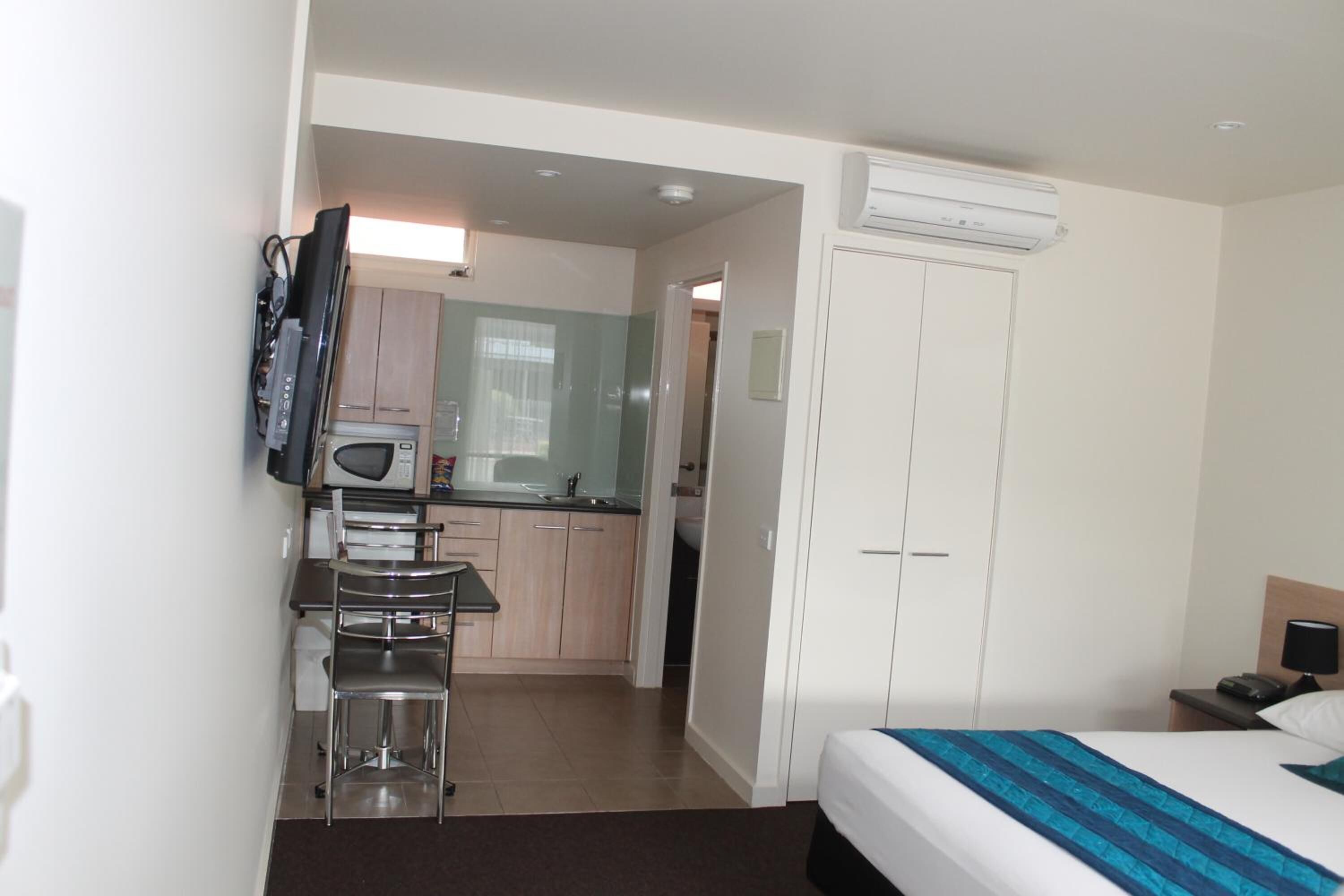 Quarto de hotel com cama, escrivaninha, cadeira, micro-ondas, pequena cozinha e guarda-roupa.