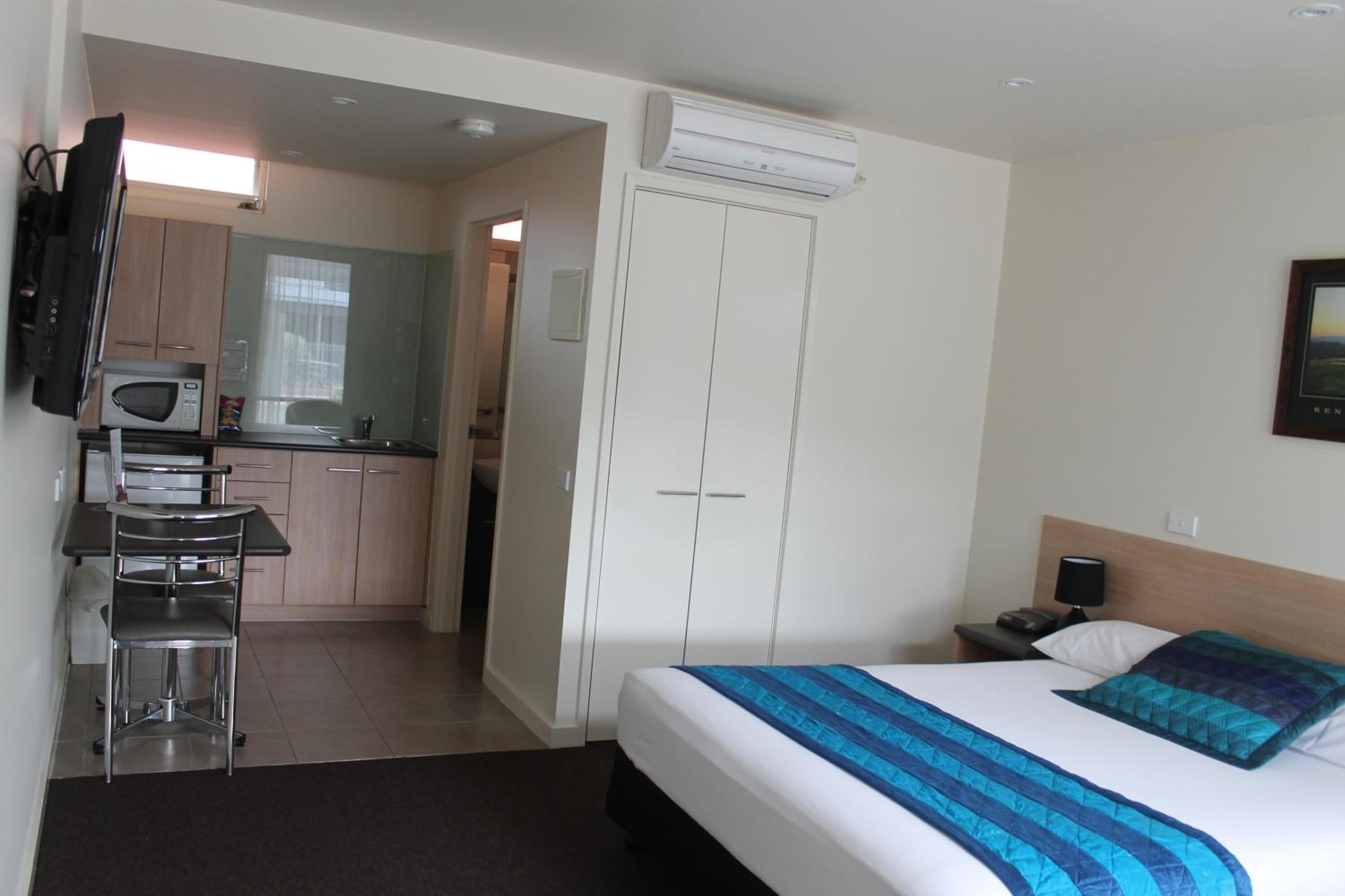 A hotel room with a bed, a desk, a chair, a microwave, a wardrobe, and an air conditioning unit.