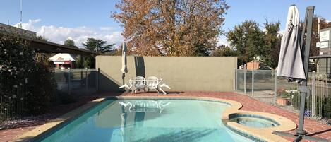 A swimming pool with a tiled edge, surrounded by a brick patio. There are two white chairs and a table, and a tree with autumn leaves in the background.