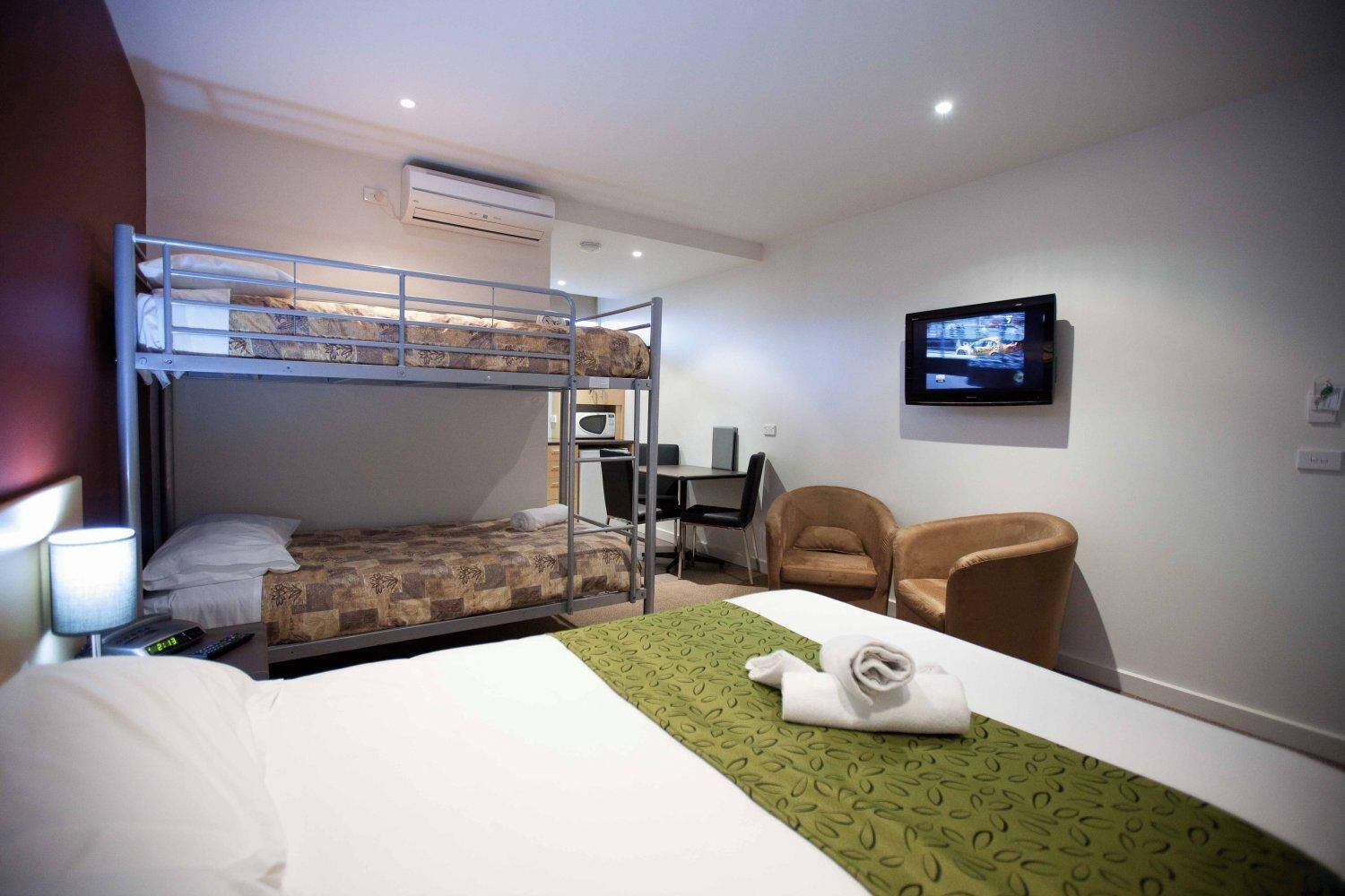 A hotel room with bunk beds, a desk, a chair, a TV, and a bed with a green bedspread.