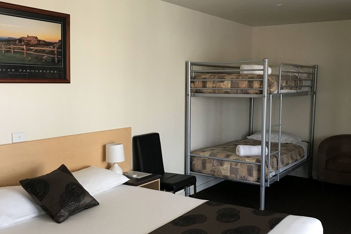 A hotel room with a bunk bed, a single bed, a headboard, a bedside table with a lamp, a chair, and a framed picture on the wall.