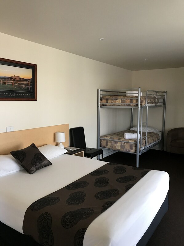 A hotel room with a bunk bed, a single bed, a headboard, a bedside table with a lamp, a chair, and a framed picture on the wall. - Mansfield Motel (Mansfield)