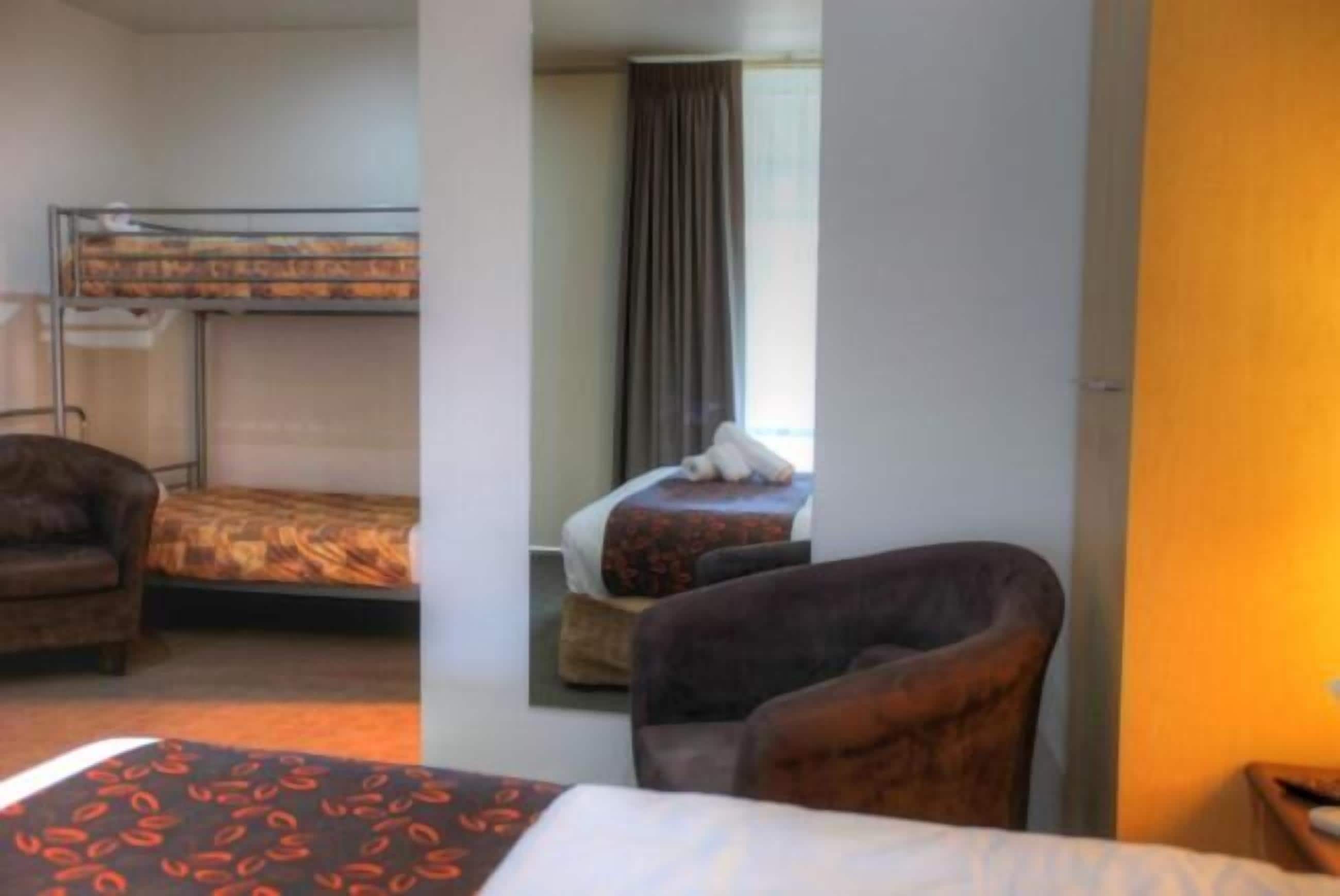 A hotel room with a bunk bed, a single bed, a chair, and a window with curtains.