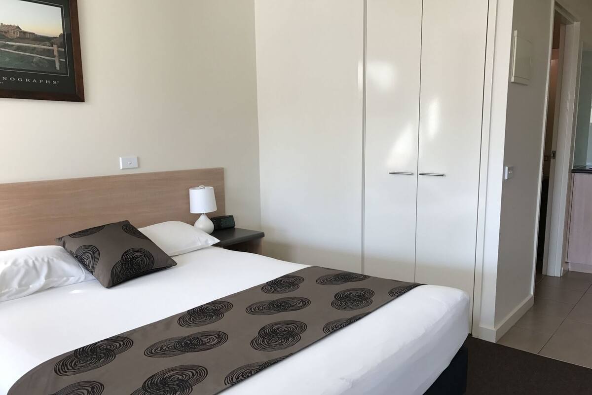 A hotel room with a bed, a nightstand, a lamp, a digital clock, and a wardrobe.