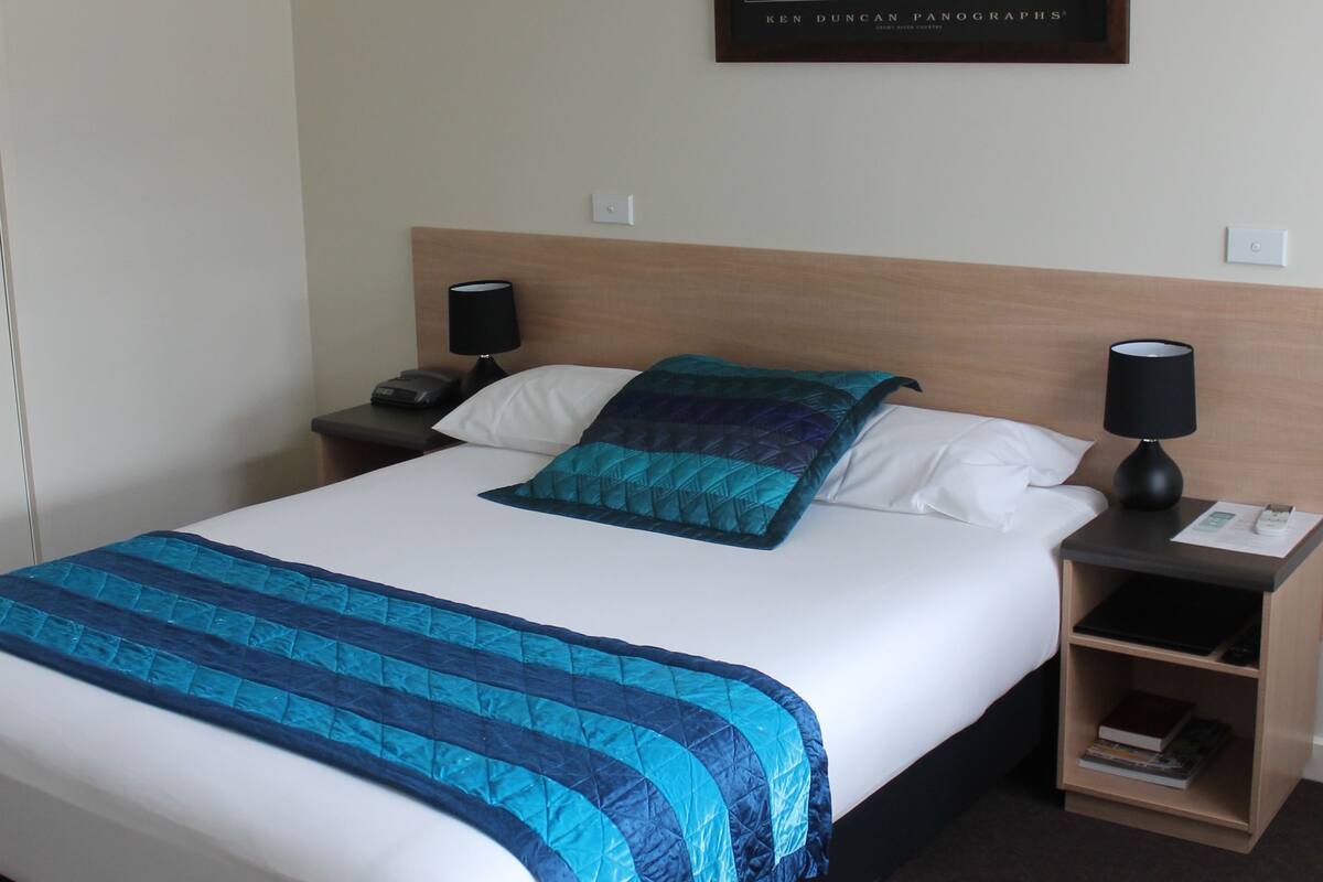A hotel room with a large bed, two bedside tables, a nightstand, a framed picture on the wall, and a carpeted floor.