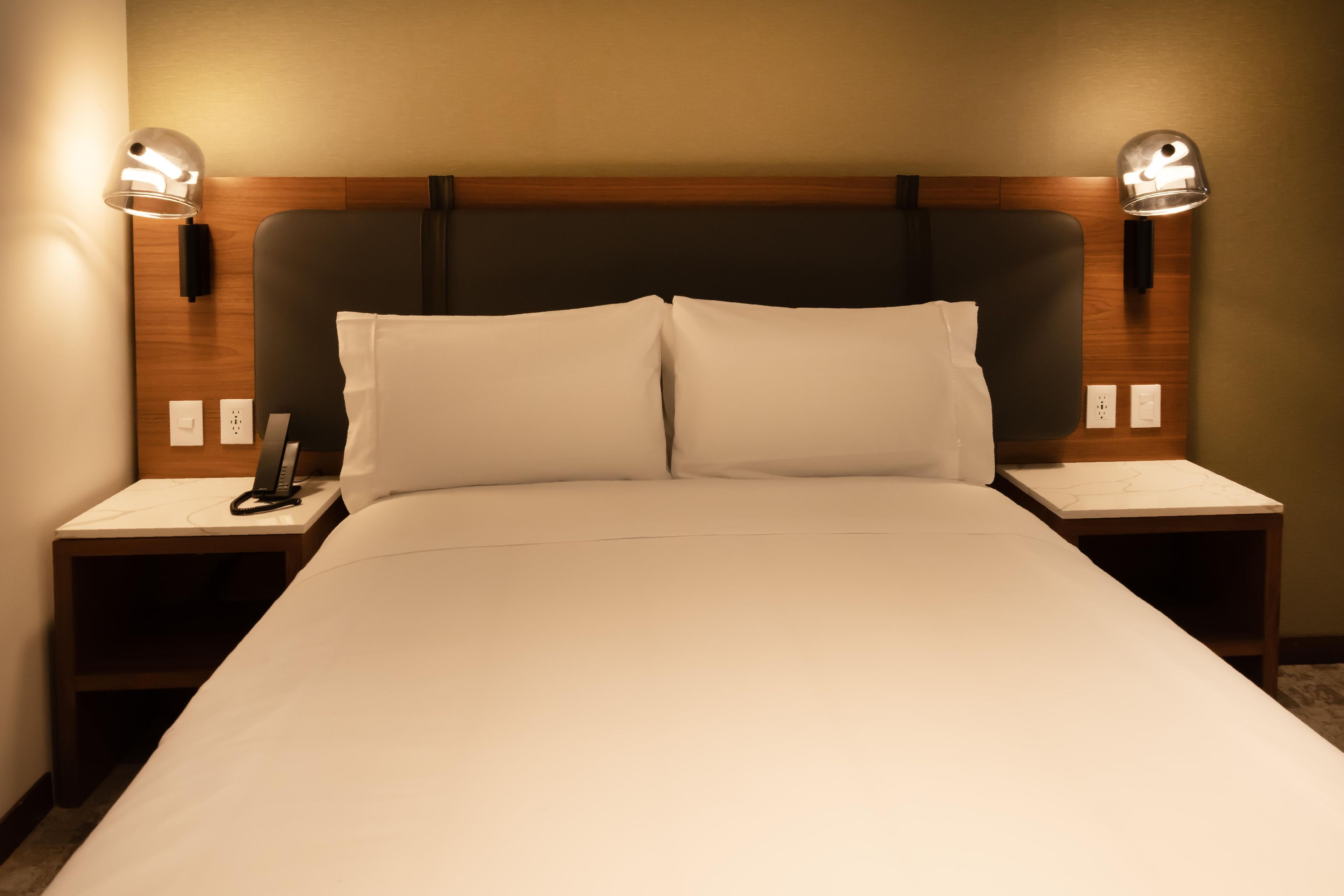 Quarto de hotel com uma cama grande, duas mesas de cabeceira, telefone e luminárias fixadas na parede.