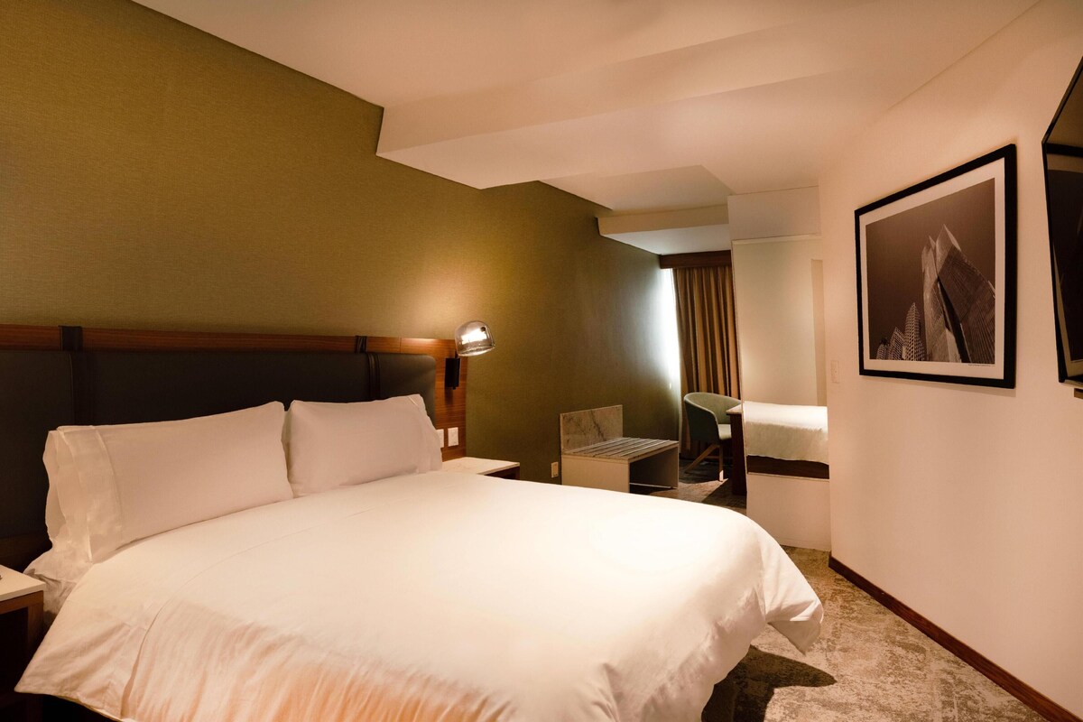 Habitación de hotel con una cama grande, un escritorio con lámpara, una silla e imagen enmarcada en la pared.