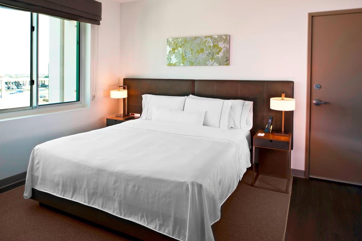 Une chambre d’hôtel avec un grand lit, deux tables de chevet avec des lampes, une fenêtre avec des stores, une porte et une œuvre d’art abstraite accrochée au mur.