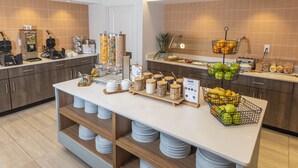 A hotel kitchen with a buffet setup, including a coffee machine, fruit baskets, and a variety of food items.