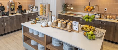 Cozinha de hotel com buffet, incluindo máquina de café, cestas de fruta e diversas opções de comida.