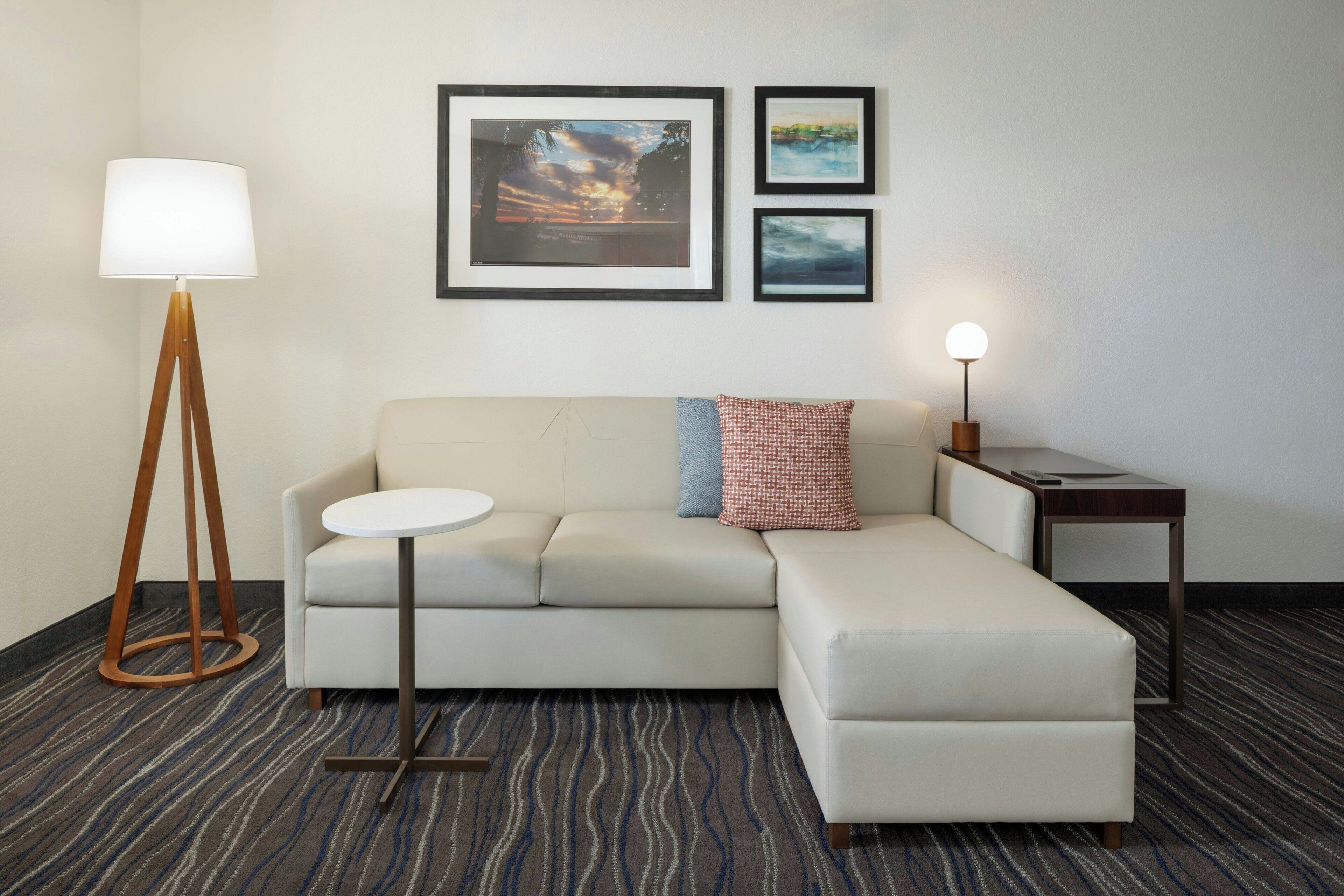 A modern hotel room with a sectional sofa, a round table, a floor lamp, and two framed artworks on the wall.