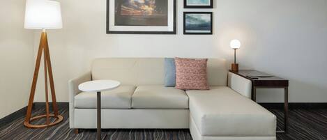Quarto de hotel moderno com sofá modular, uma mesa redonda, abajur e duas obras de arte emolduradas na parede.