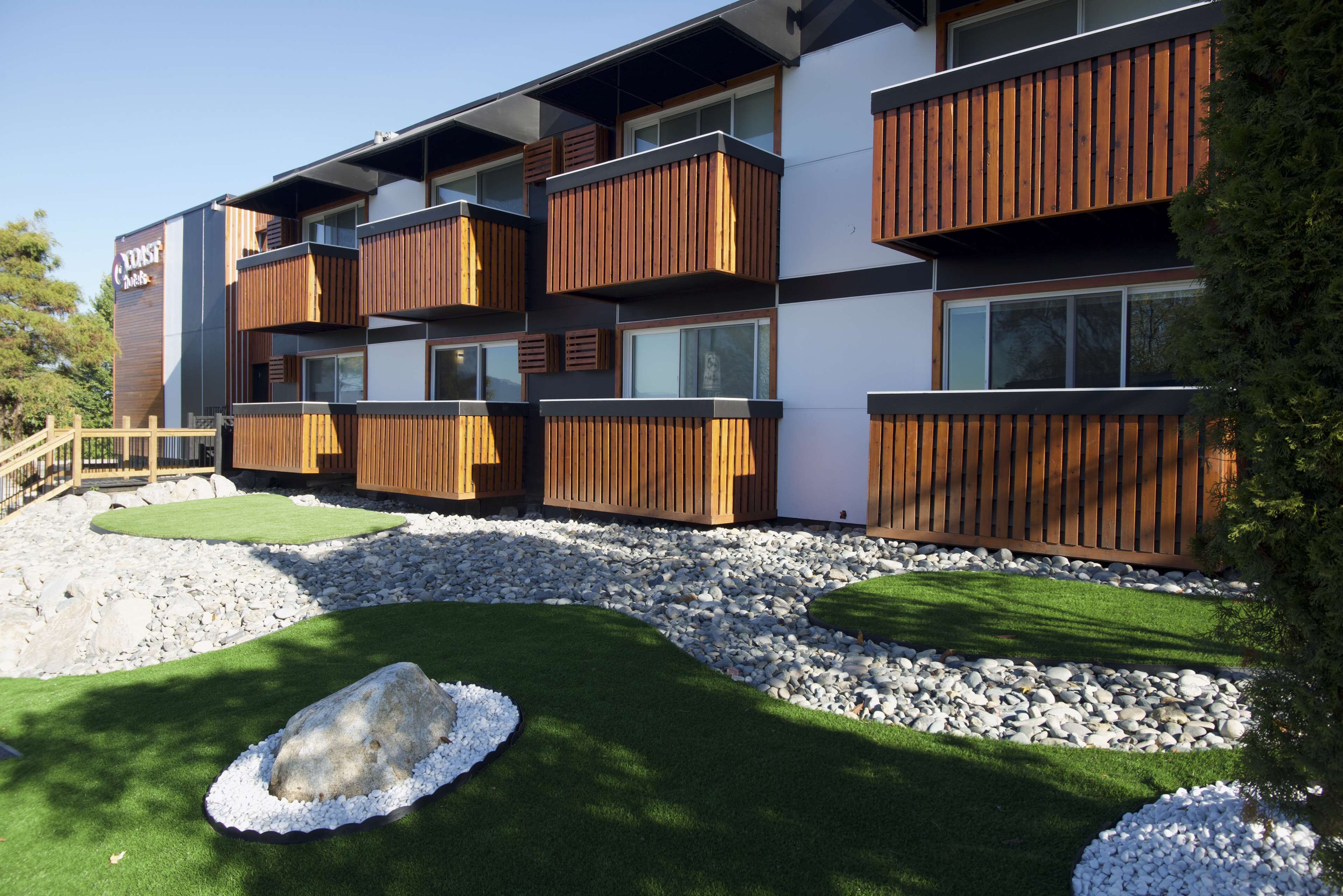 A modern building with multiple balconies, a landscaped garden with artificial grass and rocks, and a wooden fence.