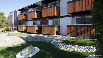 Un edificio moderno con varios balcones, un jardín ajardinado con césped artificial y rocas, y una valla de madera.