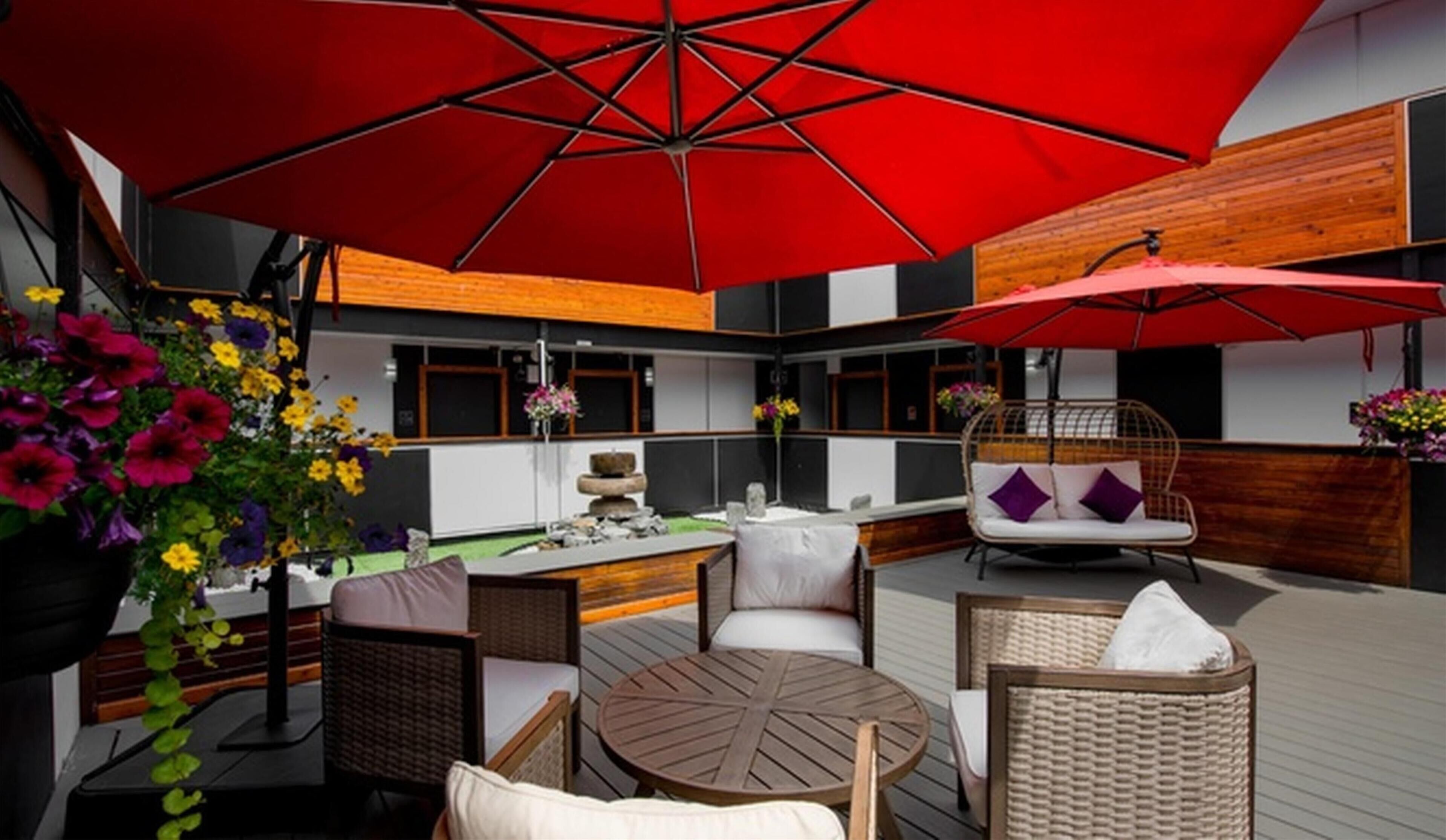 A modern outdoor seating area with wicker furniture, a round wooden table, and red umbrellas providing shade.
