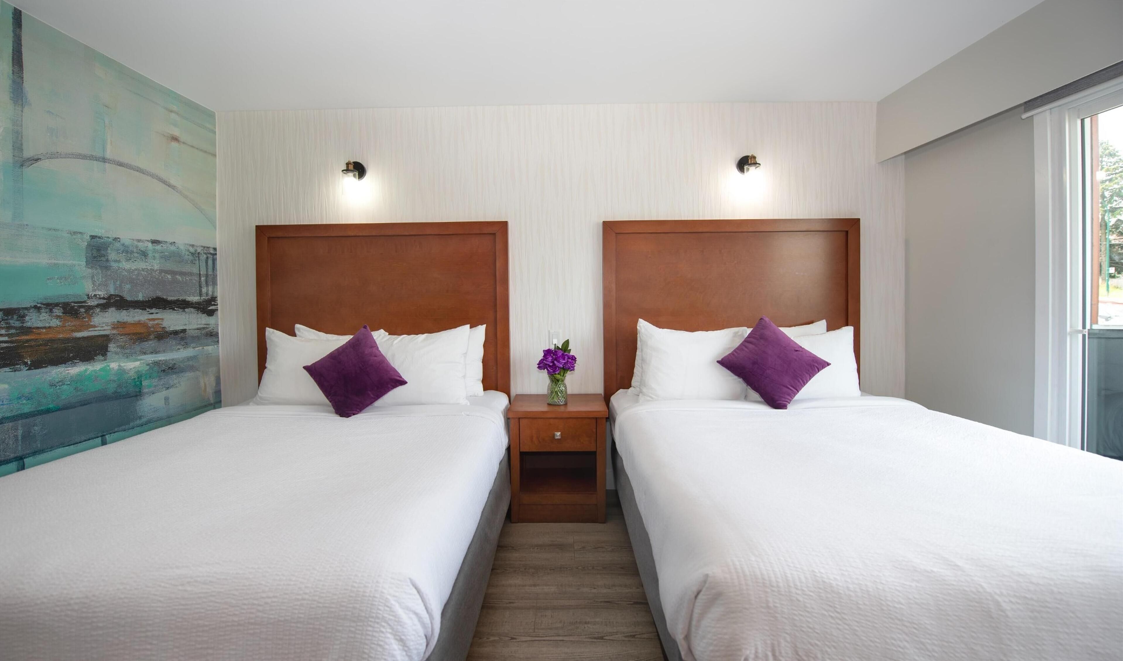 Habitación de hotel con dos camas, una mesita de noche con un florero y un cuadro abstracto grande en la pared izquierda.