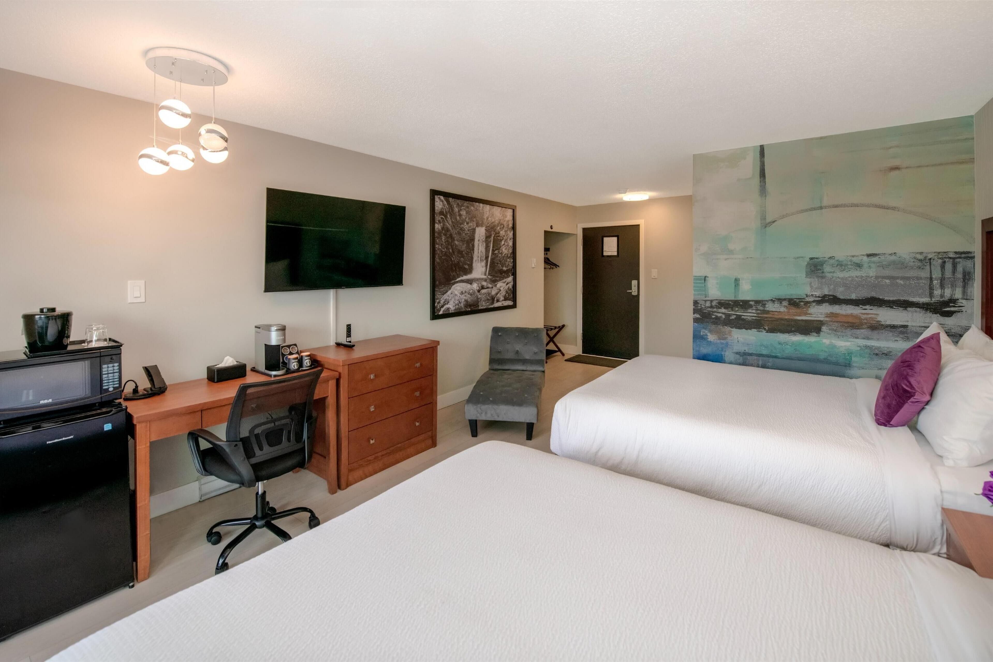 A hotel room with two beds, a desk with a computer, a chair, a TV, and artwork on the walls.