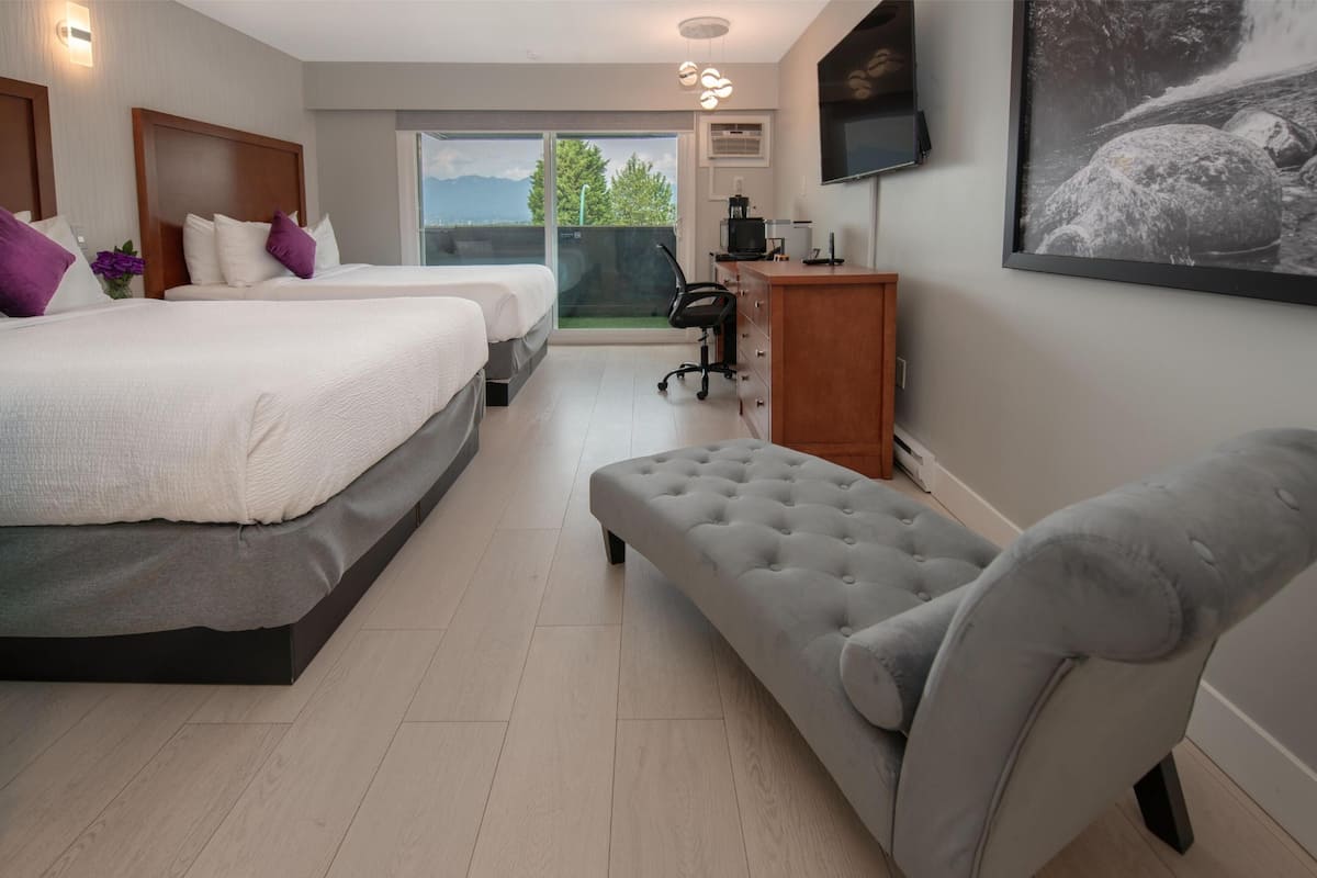 Una habitación de hotel moderna con dos camas, un sillón gris, un escritorio con computadora y un amplio ventanal con vistas a zonas verdes.