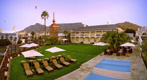 A pool area with lounge chairs and umbrellas, a stone tower, and a view of a cityscape and mountains in the background. - Dock House Boutique Hotel & Spa by NEWMARK (Cape Town)