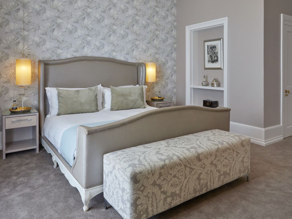 Une chambre à coucher avec un grand lit, deux tables de chevet, un banc et un mur orné d’un motif floral.