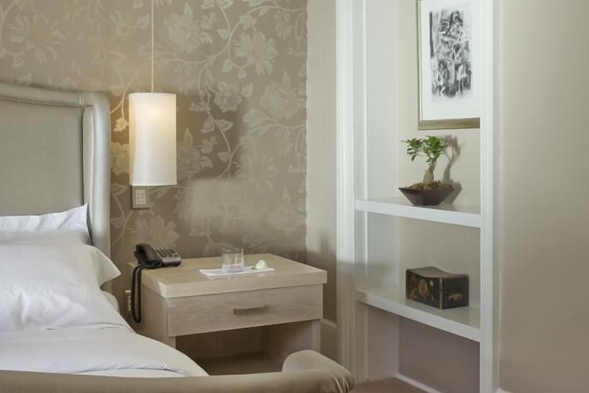 Un lit bien fait, avec du linge de lit blanc, une lampe de chevet, un téléphone et une petite table sur laquelle se trouvent un verre et un bloc-notes.