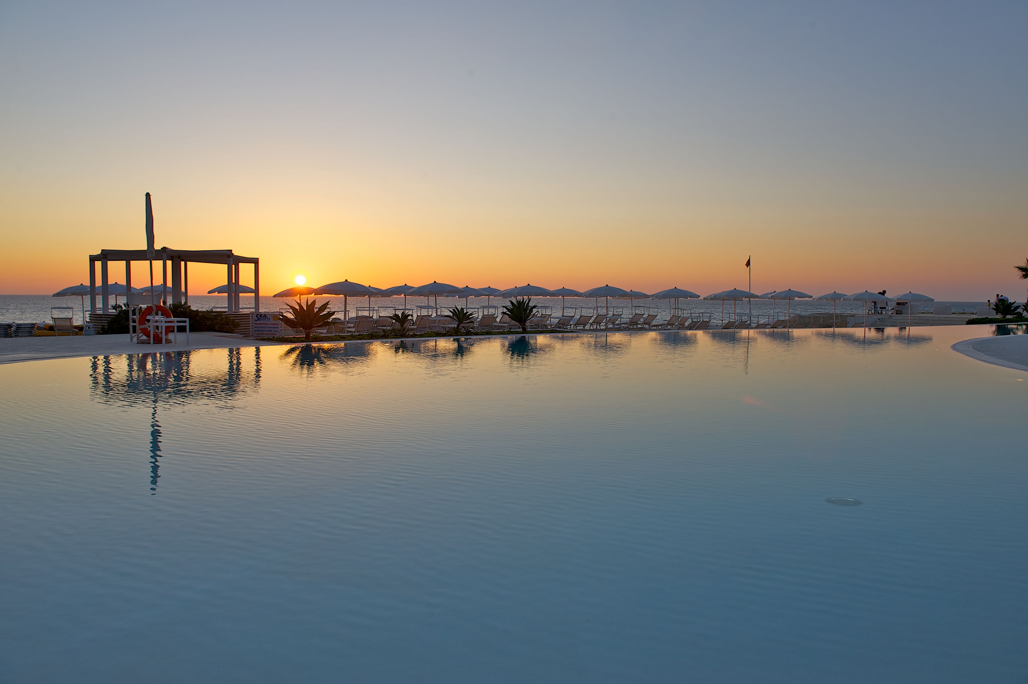 Ein friedliches Schwimmbecken mit Liegestühlen und Sonnenschirmen, umgeben von Bergen und einem Sonnenuntergang.