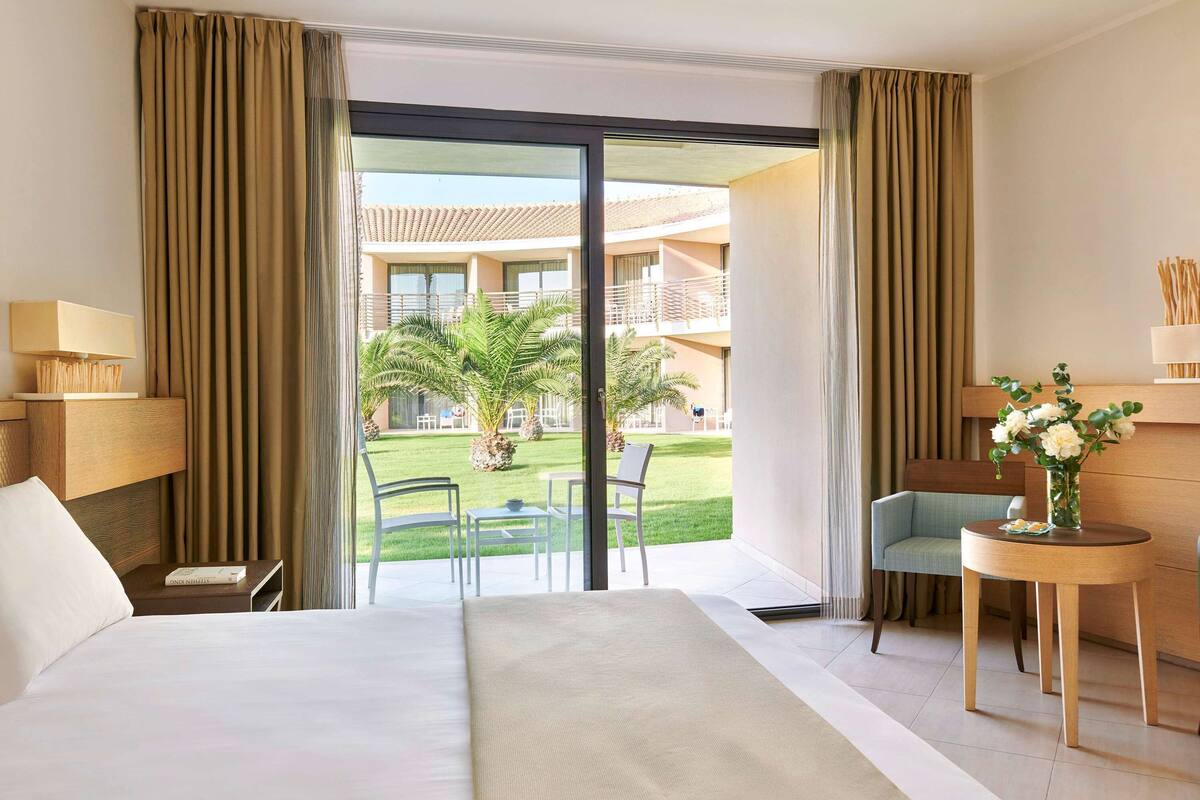 Habitación de hotel con cama, escritorio, silla y vista a un patio con palmeras y un edificio al fondo.