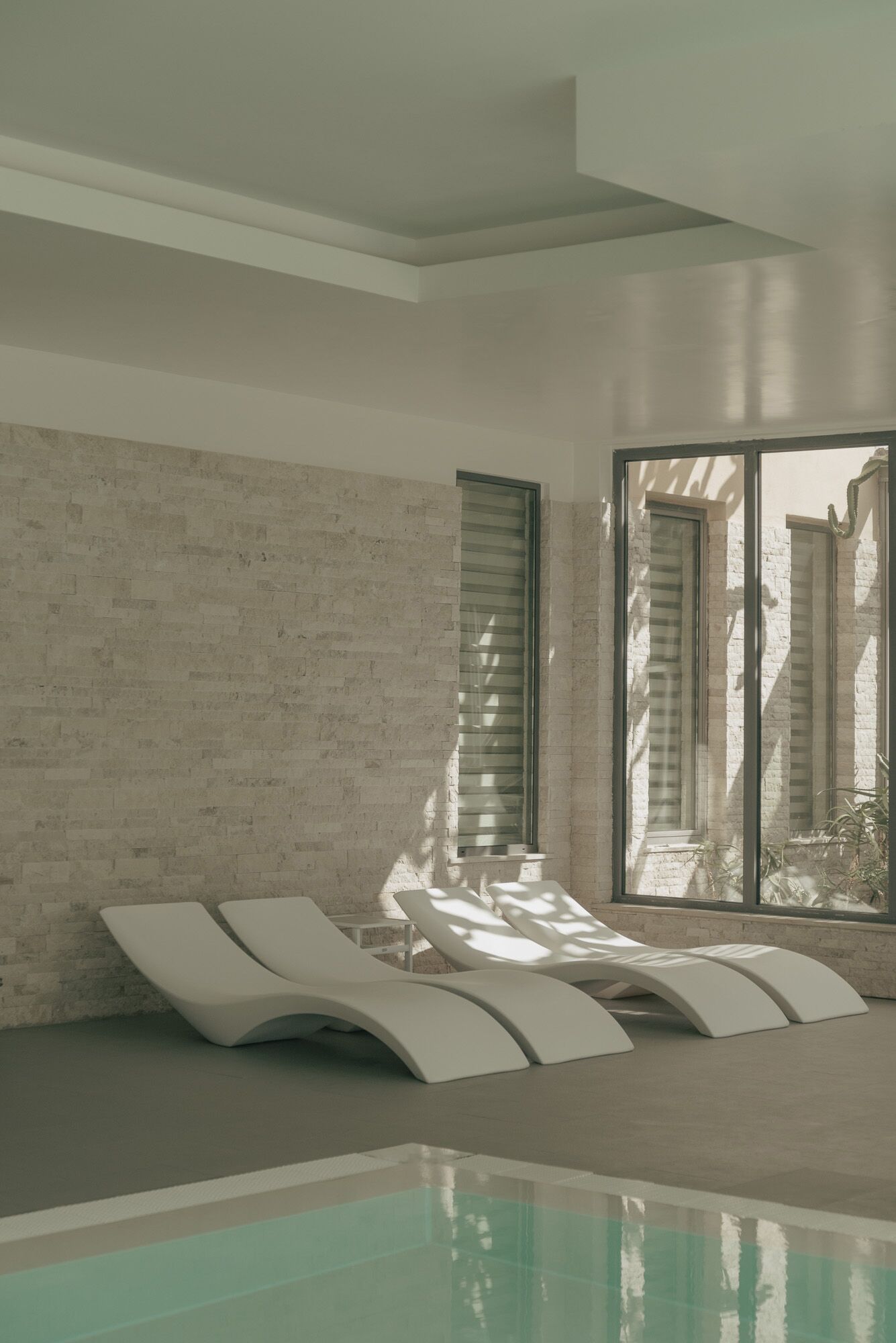 Un área de piscina interior moderna con pared de ladrillo, ventanas amplias y sillones blancos.