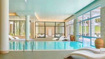 Piscina interior moderna con paredes de vidrio transparente, sillones blancos y vistas al exterior.
