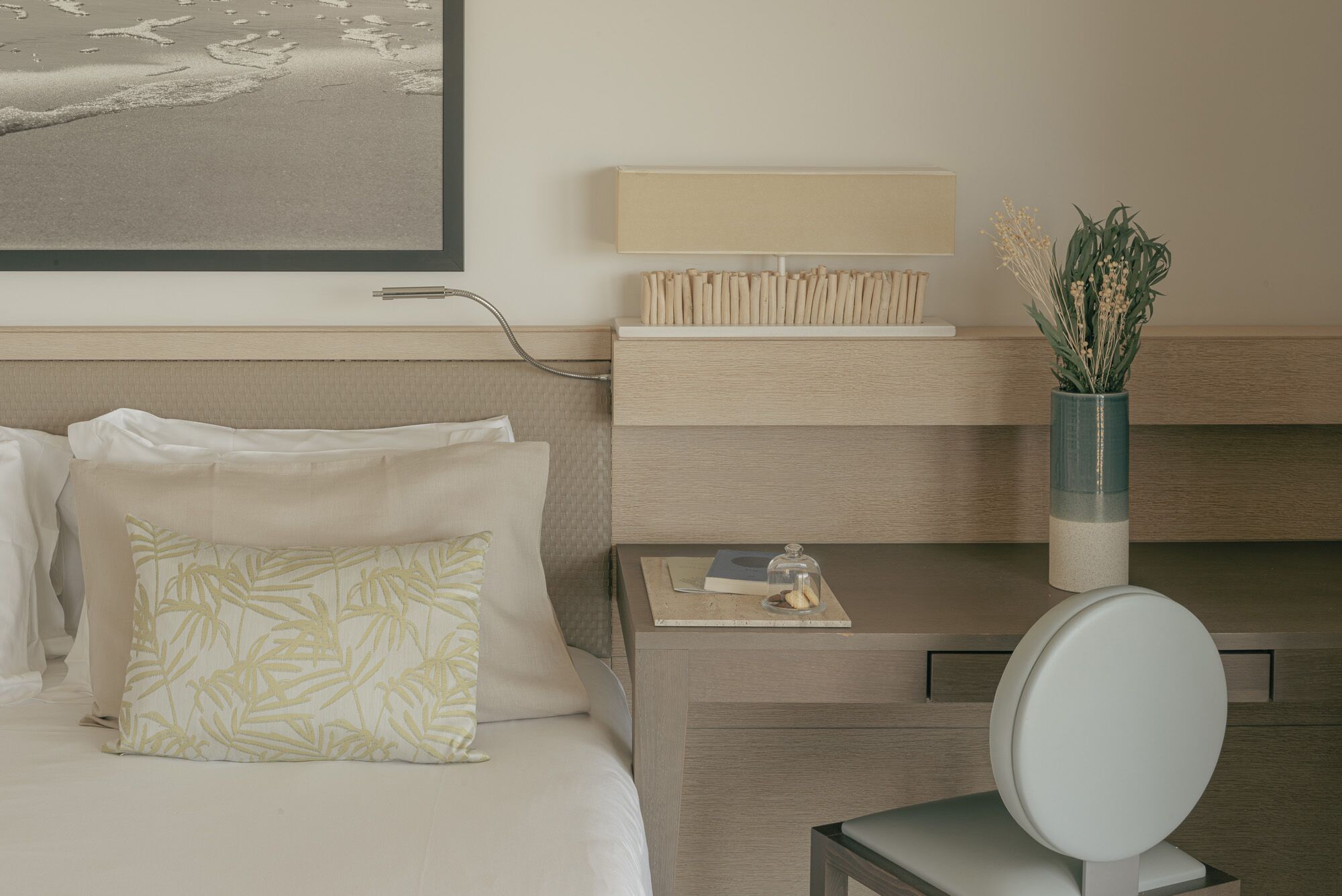 A hotel room with a bed, a bedside table with a lamp, a vase with flowers, and a framed picture on the wall.