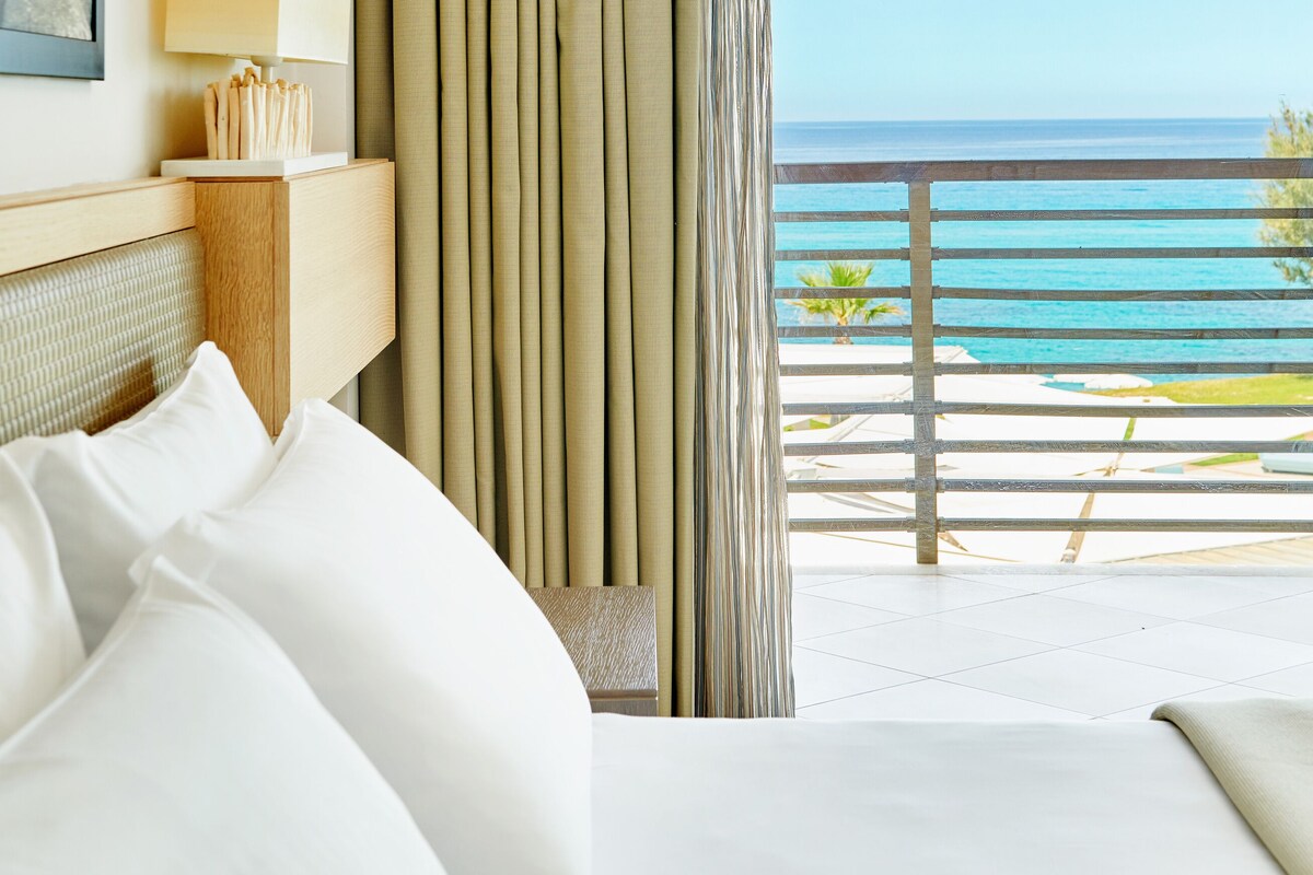 A hotel room with a bed, a nightstand with a lamp, a view of the ocean, and a balcony with a railing.