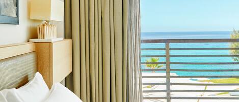 A hotel room with a bed, a nightstand with a lamp, a view of the ocean, and a balcony with a railing.