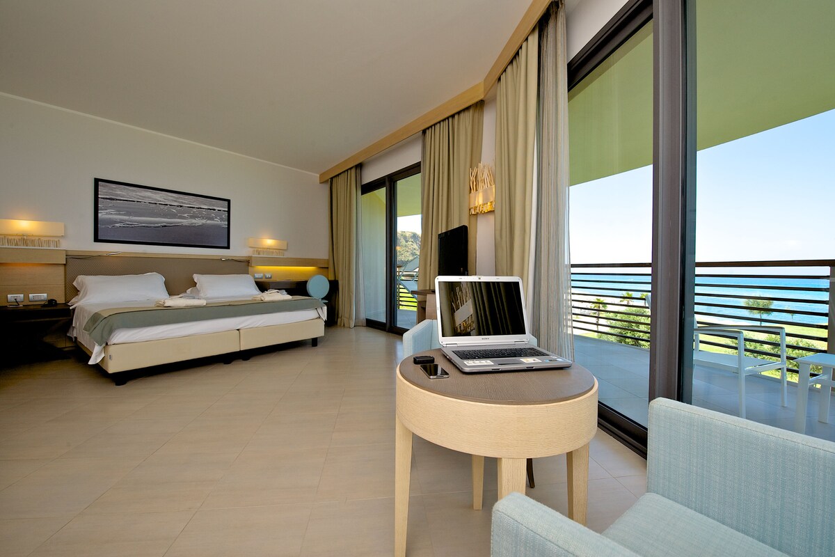 A modern hotel room with a bed, a desk with a laptop, a chair, and a balcony with a view.