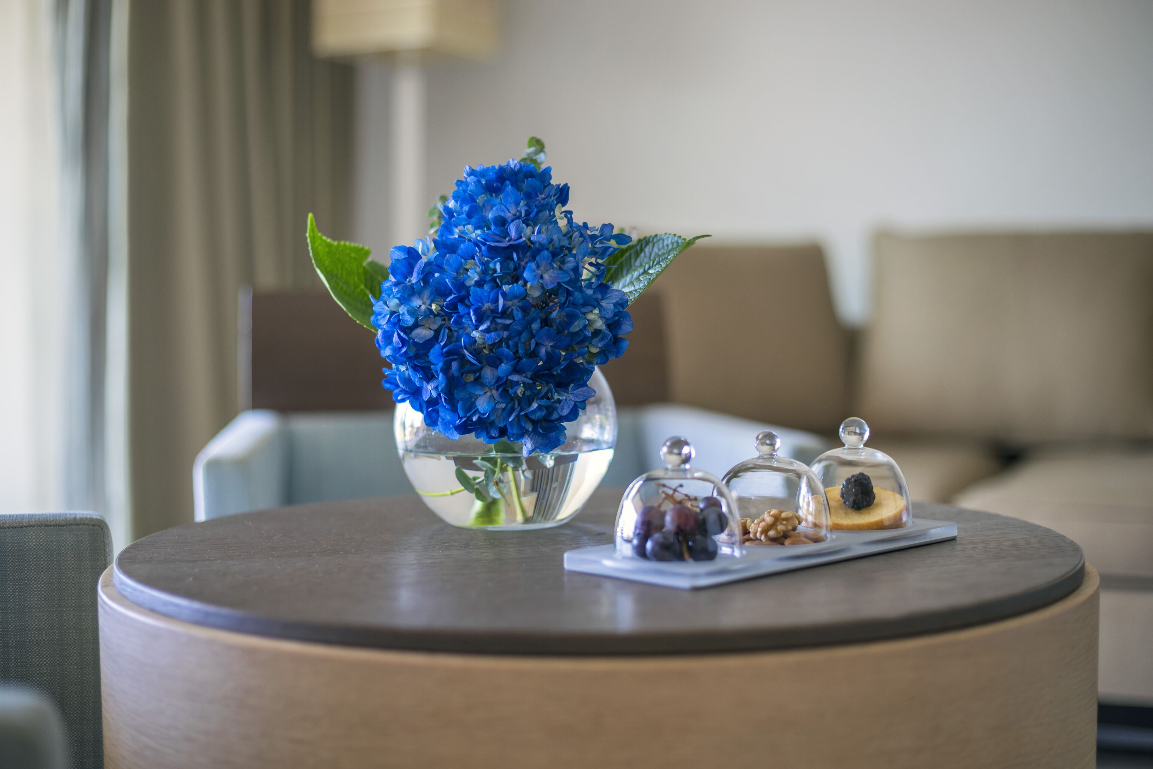 A round wooden table with a vase of blue flowers, a glass dome with a fruit and nuts, and a small plate with a piece of bread.
