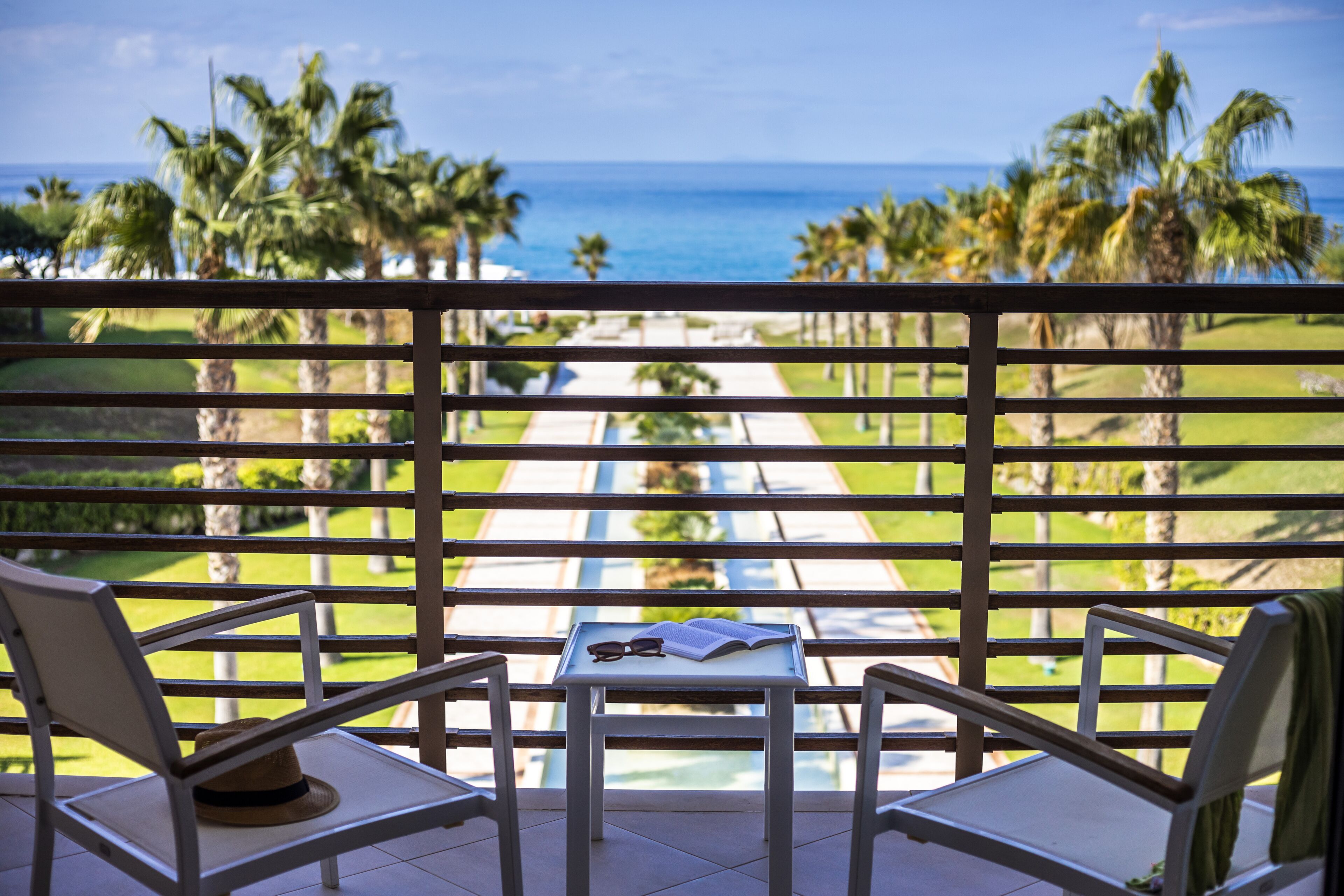 A balcony with a view of the ocean, palm trees, and a table with a hat and glasses.