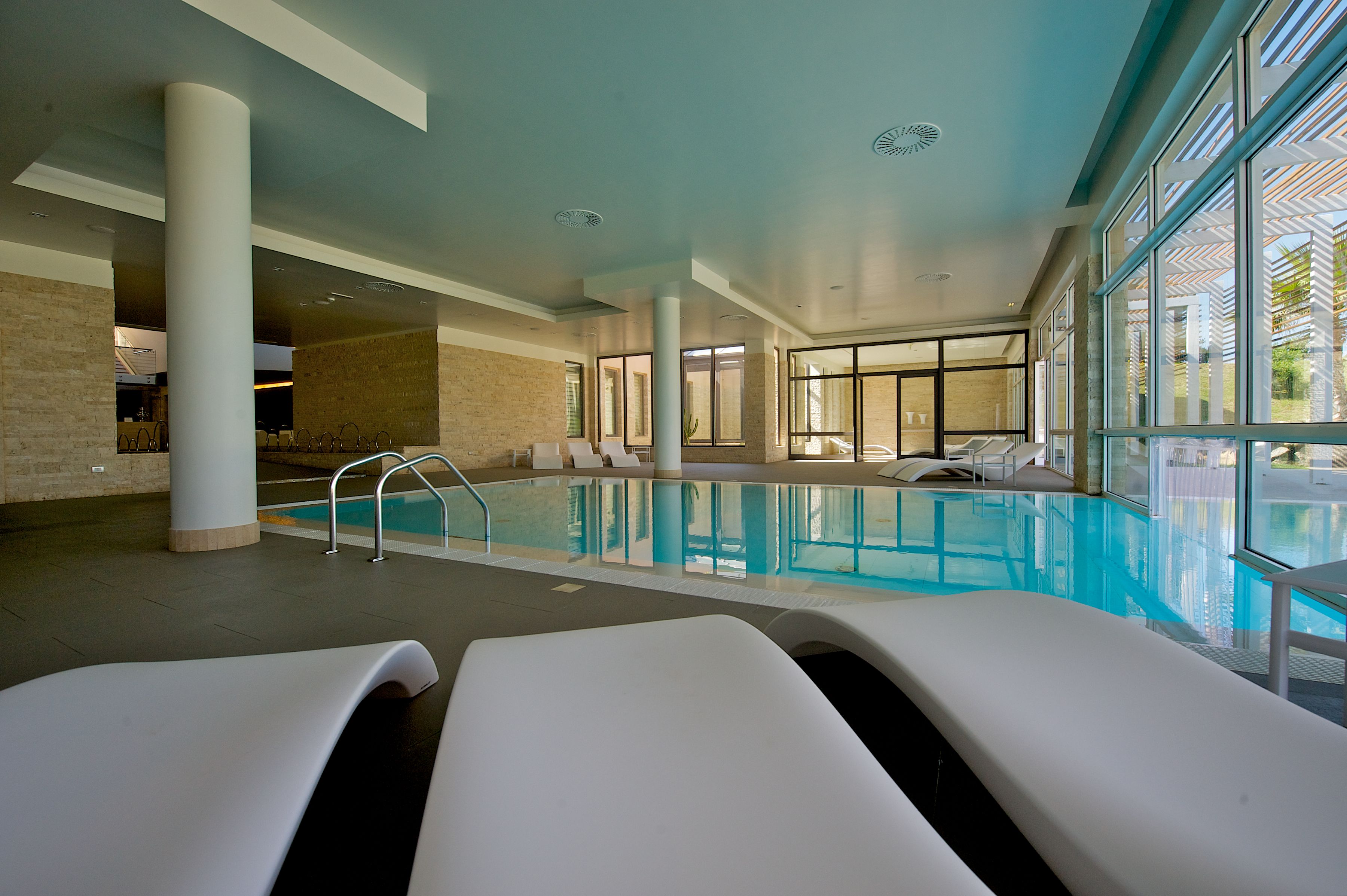 A modern indoor swimming pool with clear glass walls, a poolside lounge area, and a view of the outdoors.