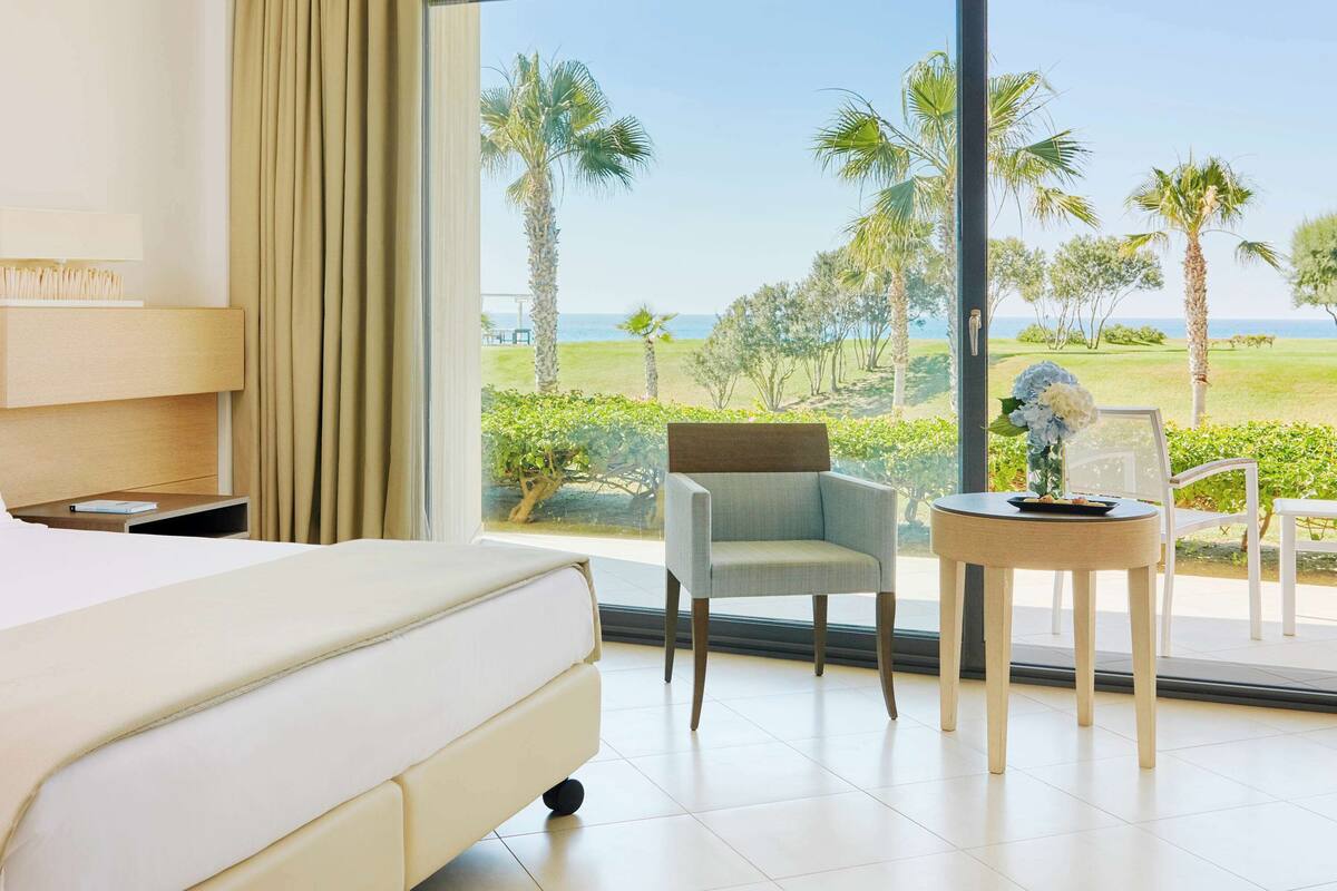 Habitación de hotel con una cama, una silla, una mesita pequeña y un ventanal con vistas a la playa y palmeras.