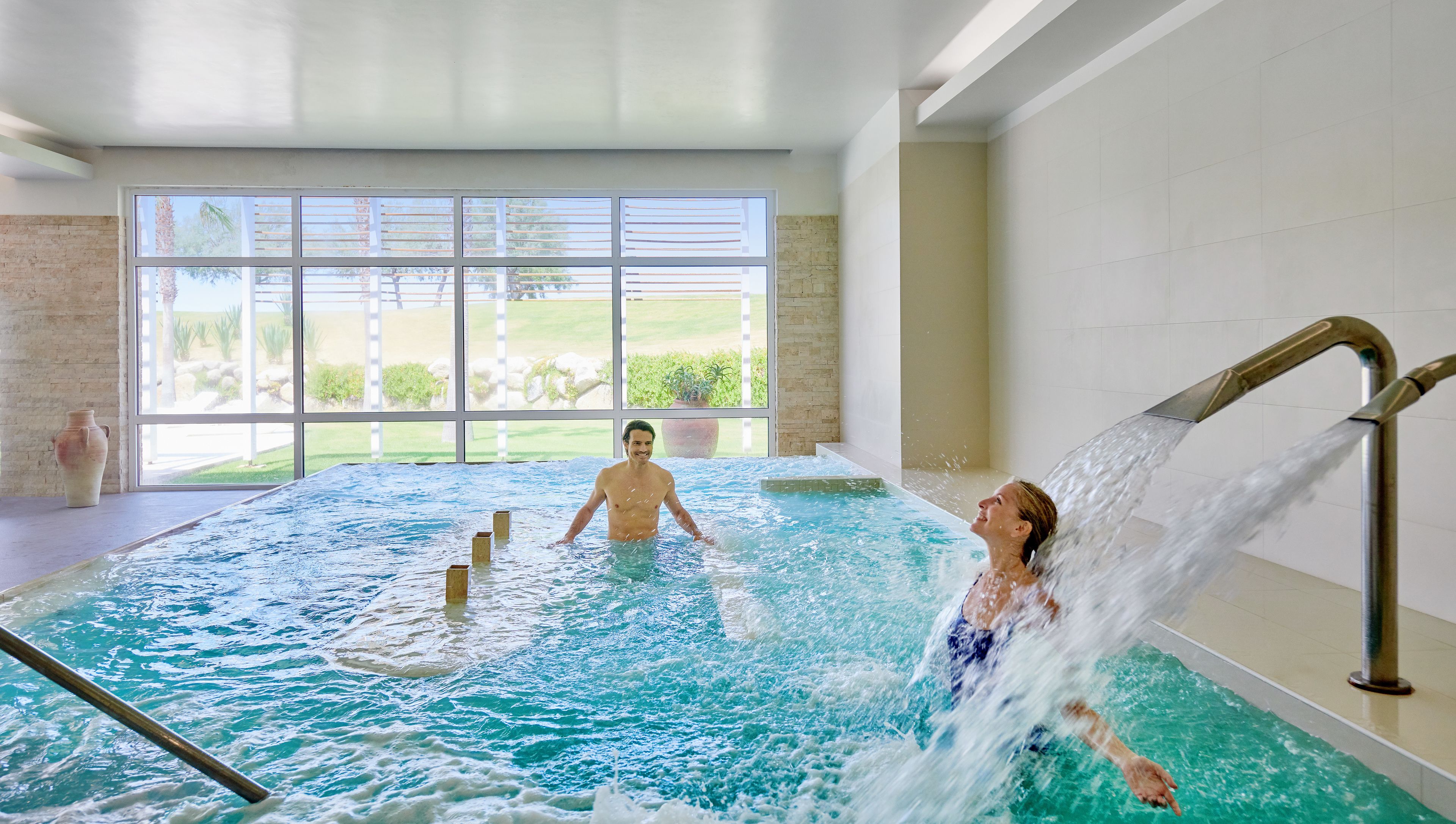 Una piscina interior con agua cristalina, una persona de pie en la piscina y otra persona bajo un cabezal de ducha.