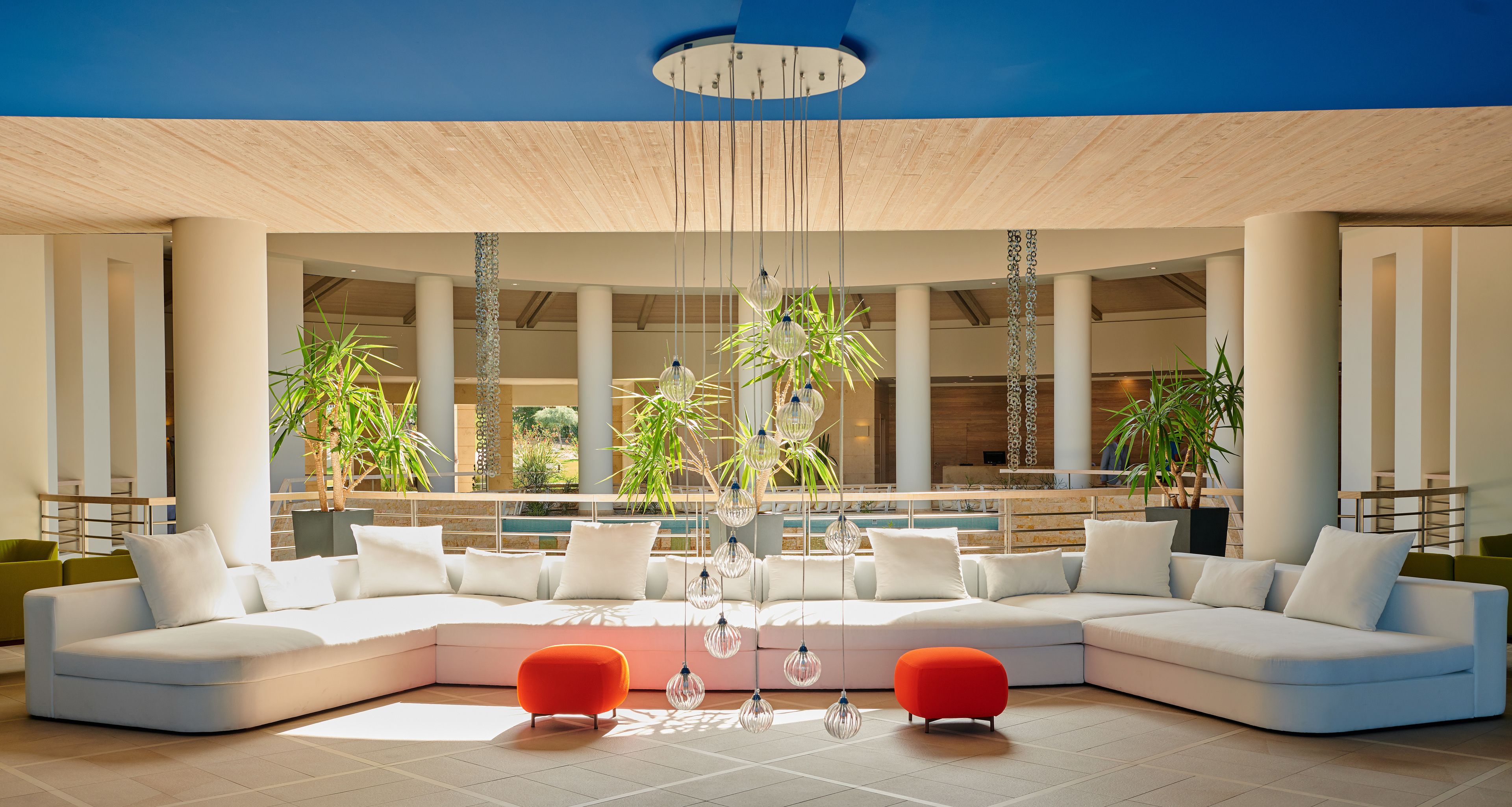 Un hall d’entrée moderne avec un canapé d’angle, un pouf orange, des luminaires suspendus en verre et de grandes fenêtres donnant sur un espace piscine.