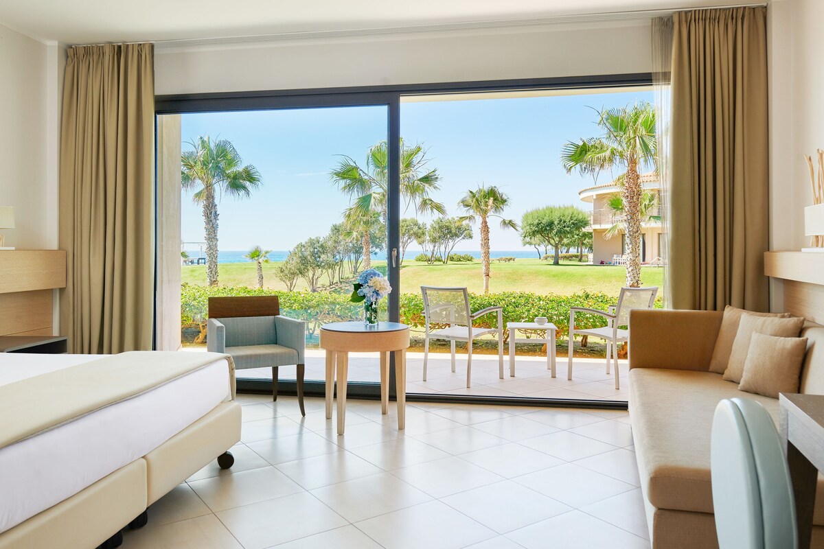 A hotel room with a bed, a sofa, a table, and chairs. There is a large window with a view of palm trees and a building.